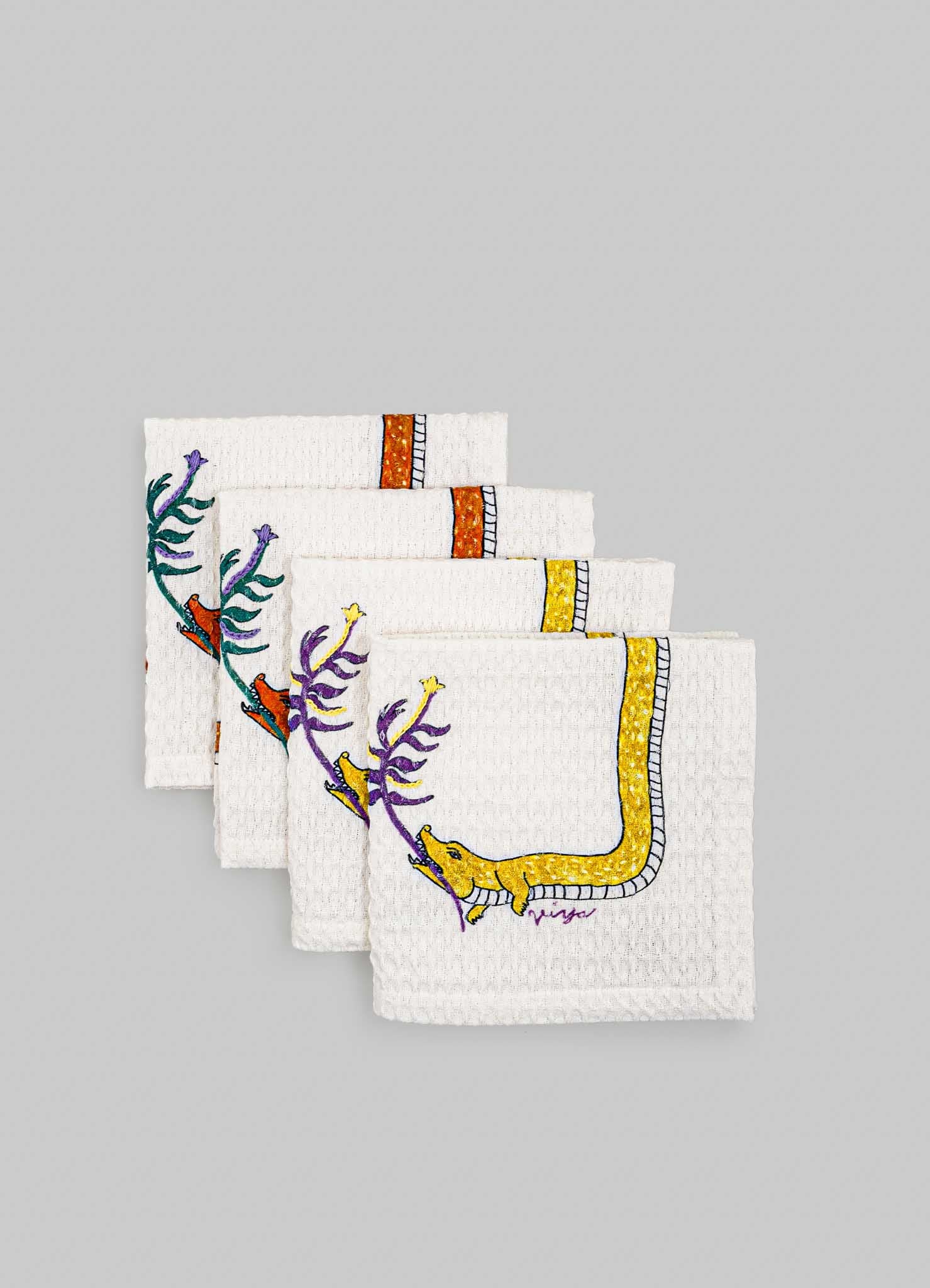 Set of white textured cloth napkins with colorful embroidered motifs of animals and plants, featuring purple, yellow, orange, and green thread details. The napkins have decorative borders and a playful, handcrafted style suitable for casual or themed dining settings.