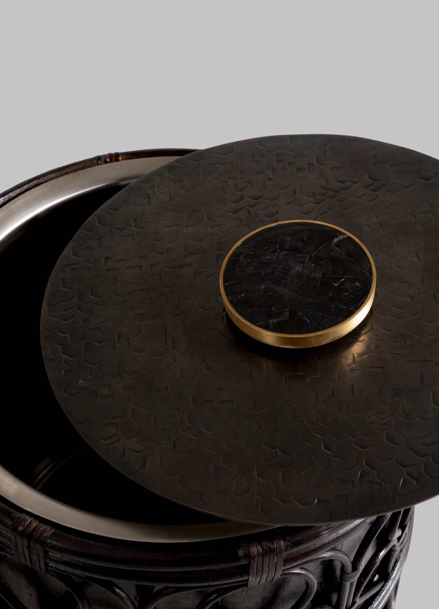A close-up of the VIYA Manipur Ice Bucket Small shows its round, textured dark metal lid with a hand-beaten brass edge, partially covering the ornate container. The intricate rim pattern stands out against a plain gray background.