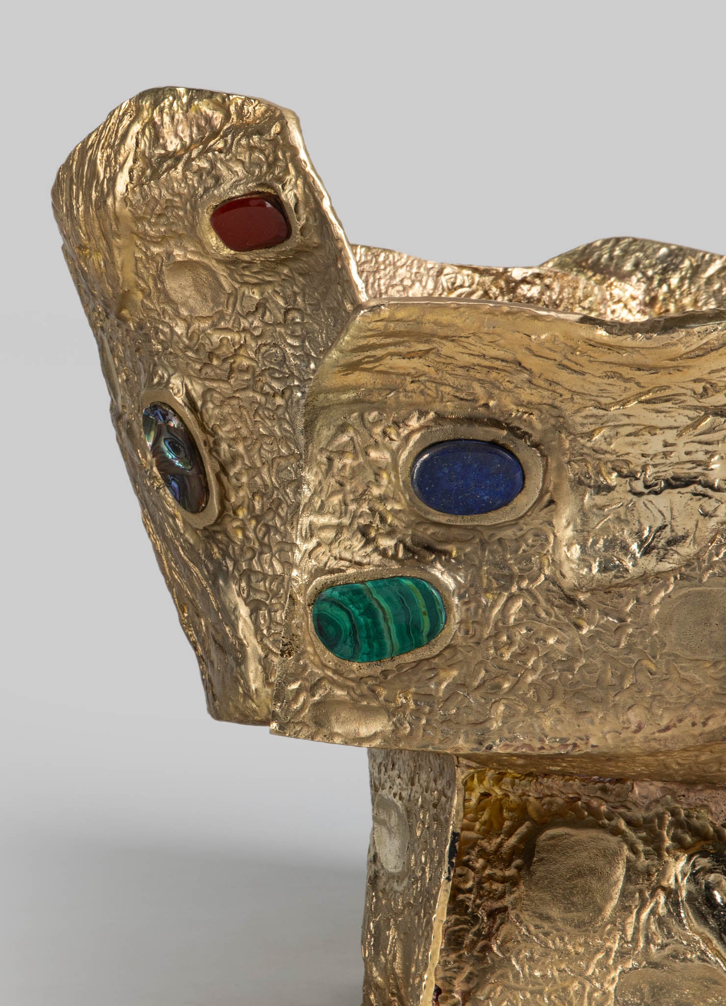 A textured gold-colored sculpture with an abstract, organic form decorated with oval-shaped inlays in red, blue, green, and iridescent hues. The surface has a rough, hammered appearance, emphasizing an artisanal, handcrafted style.
