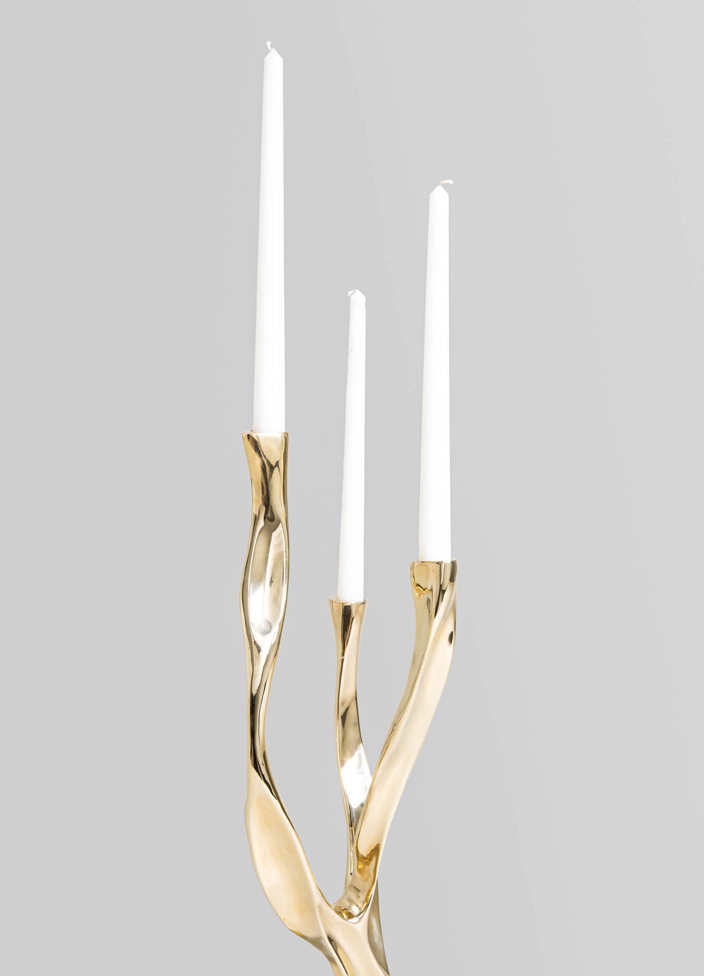 Gold-tone brass candle holder with a twisted, organic shape, holding three white tapered candles, set against a neutral gray background.