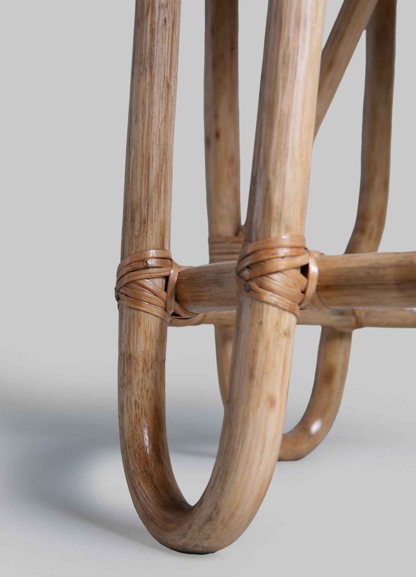 Close-up of a rattan furniture leg with a natural wood finish, featuring woven rattan bindings at the joint. The design emphasizes handcrafted craftsmanship and organic materials.