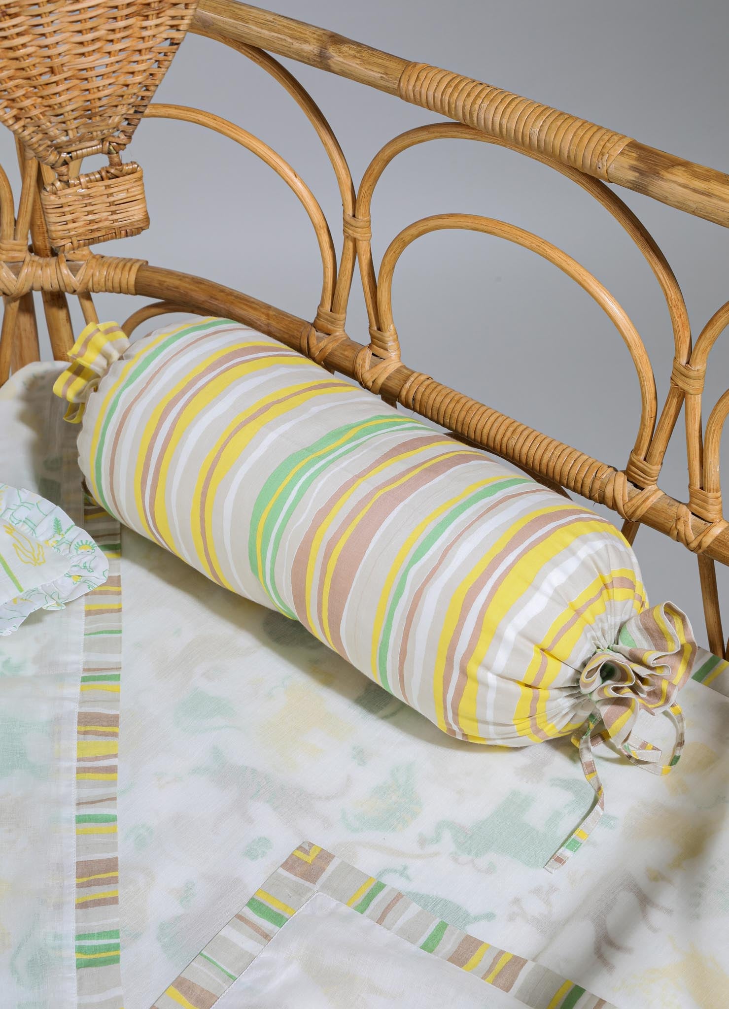 A striped fabric bolster pillow with pastel yellow, green, and beige tones, featuring fabric ties on each end, placed on a bed with a white and light-colored patterned bedsheet. The headboard is made of rattan with curved, woven details.