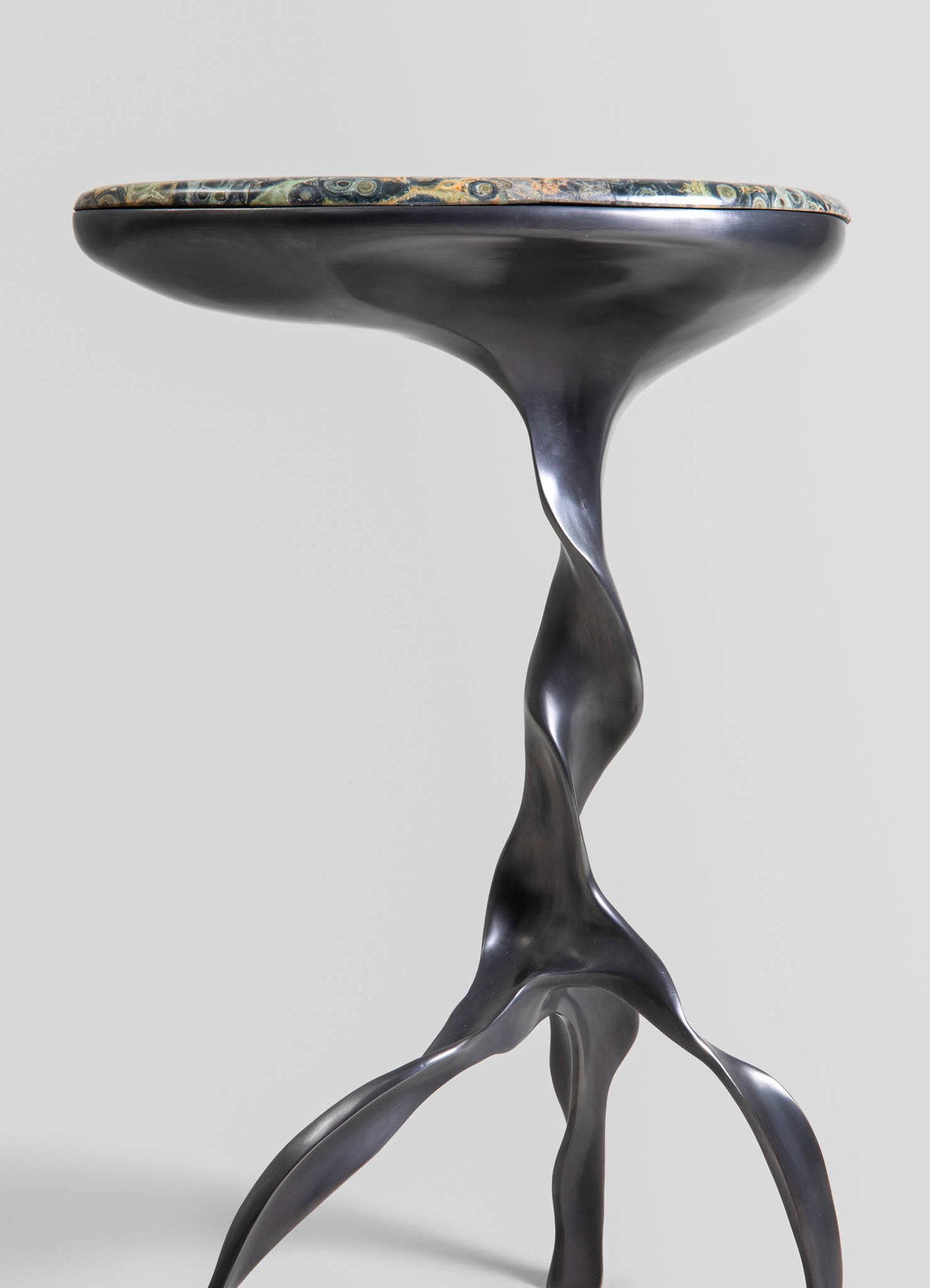 A side table with a marble top featuring swirling green, beige, and gray patterns and a sculptural, black metallic base with organic, flowing shapes.