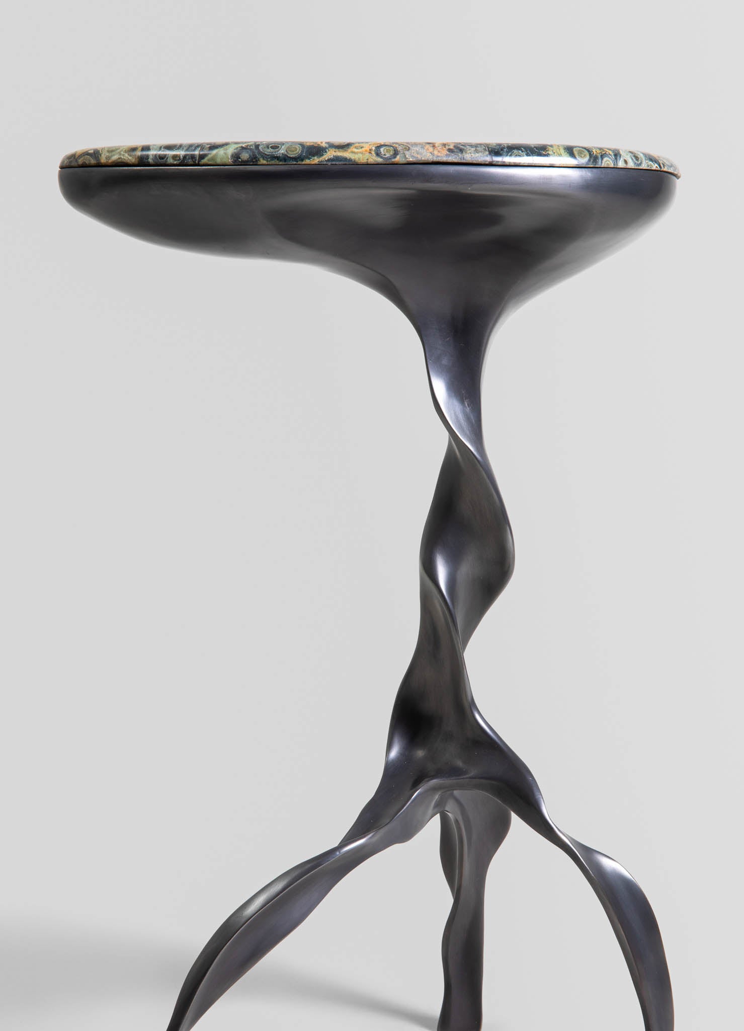 A side table with a marble top featuring swirling green, beige, and gray patterns and a sculptural, black metallic base with organic, flowing shapes.