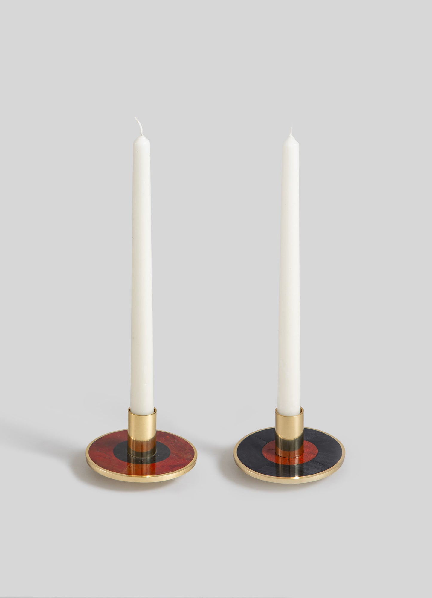 Pair of decorative candle holders with circular bases featuring geometric color blocks in red, black, and gold, with brass accents. The candle holders support tall white taper candles in a contemporary, minimalist style.