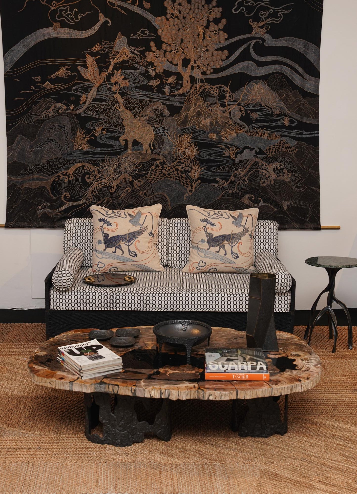 A living room interior featuring a woven jute rug, a black and white patterned sofa with silk cushion covers displaying traditional motifs, and a rustic wooden coffee table with a natural finish. The background includes a large fabric wall hanging with intricate gold and dark tones depicting a landscape scene with animals and trees.