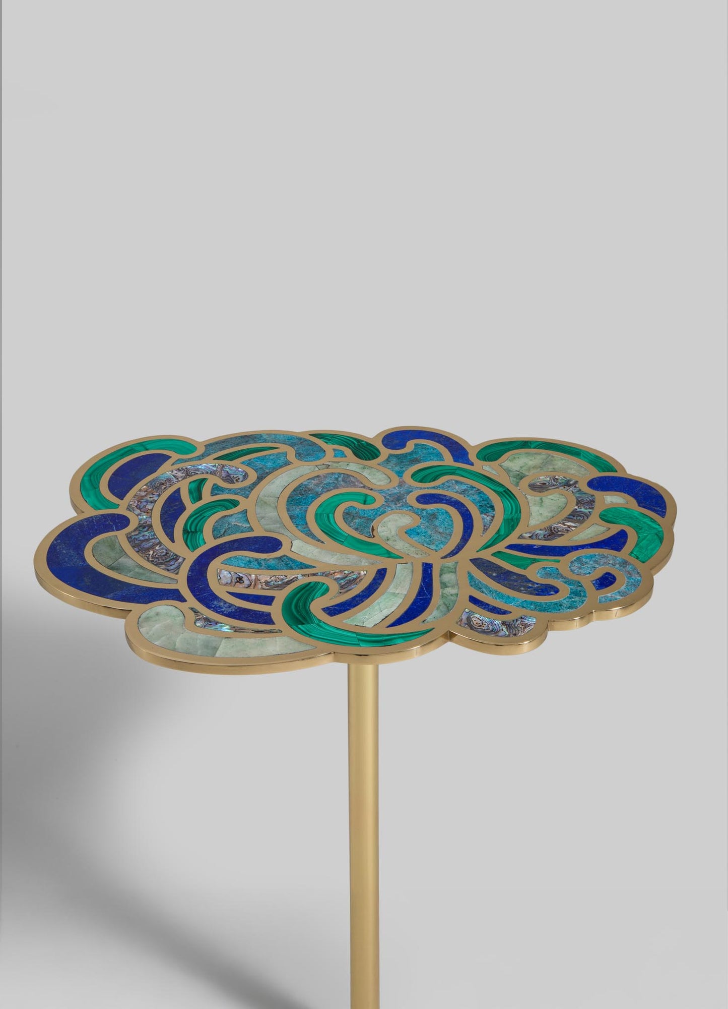 A side table with a decorative top featuring a mosaic design of swirling patterns in blues, greens, and beige, set against a plain background. The table has a slim, gold-colored metal leg.