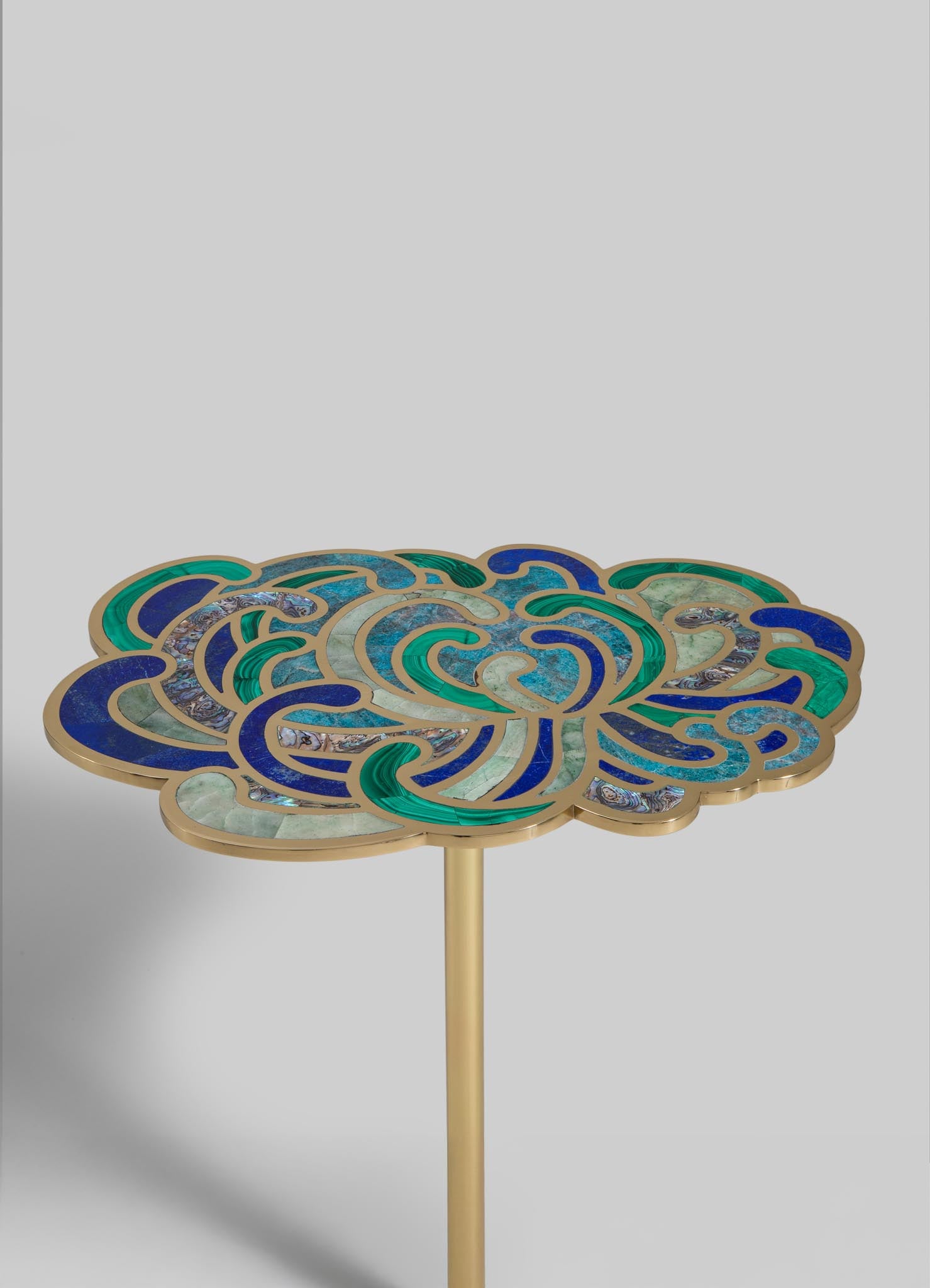 A side table with a decorative top featuring a mosaic design of swirling patterns in blues, greens, and beige, set against a plain background. The table has a slim, gold-colored metal leg.