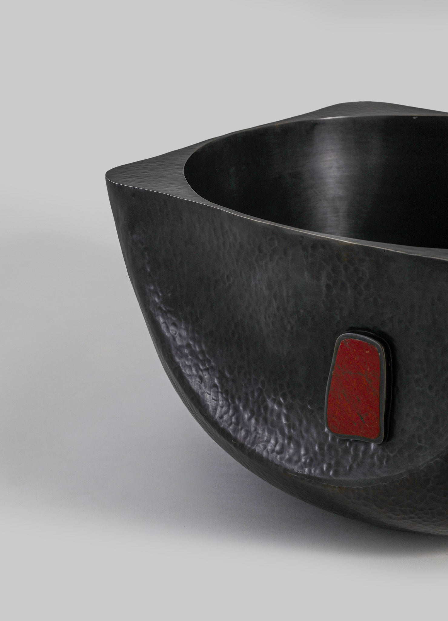 A black hammered metal bowl with a curved, organic shape and a small red rectangular detail on its exterior surface. The bowl has a contemporary design with a textured finish.