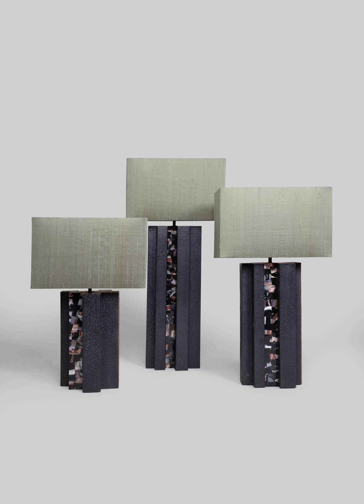 Three table lamps with rectangular fabric shades in light beige or off-white, mounted on bases with a textured black finish and decorative mosaic-like inlay in earth tones. The lamps have a modern, minimalist design with clean lines and contrasting materials.