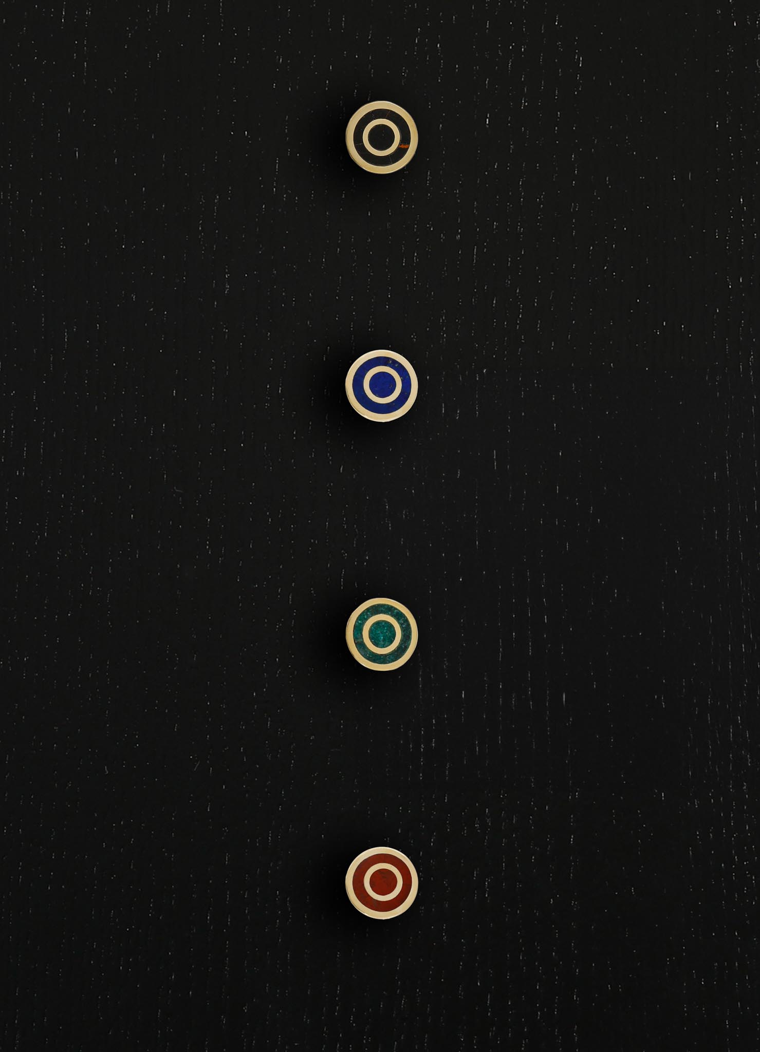 Four circular buttons with concentric ring patterns in cream, blue, green, and red, mounted on a black wooden surface. The wood has a visible grain texture, and the buttons are arranged vertically in a straight line.