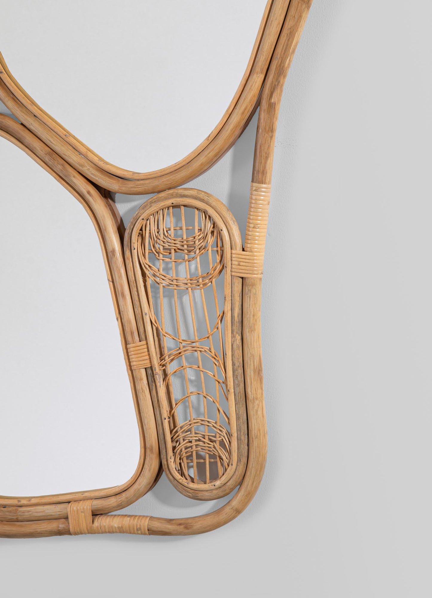 A rattan wall mirror featuring a natural wood finish and a handcrafted frame with round and linear motifs, mounted on a light-colored wall. The design incorporates woven rattan elements that add a textured, rustic style.