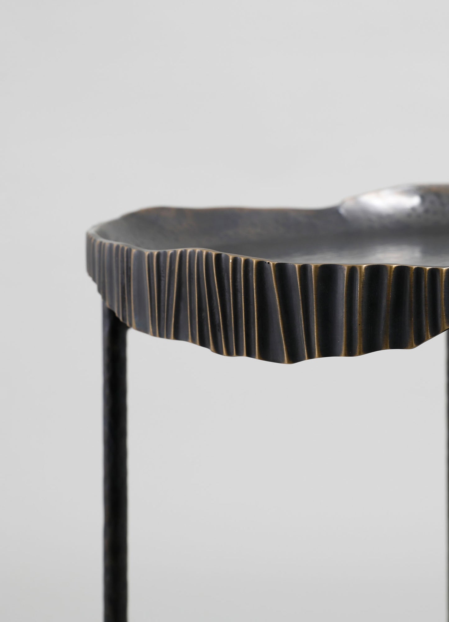A close-up of a black ceramic tray with a fluted, textured edge and a glossy finish, supported by slender metal legs. The tray has an organic, handcrafted design with subtle gold accents along the ridges.