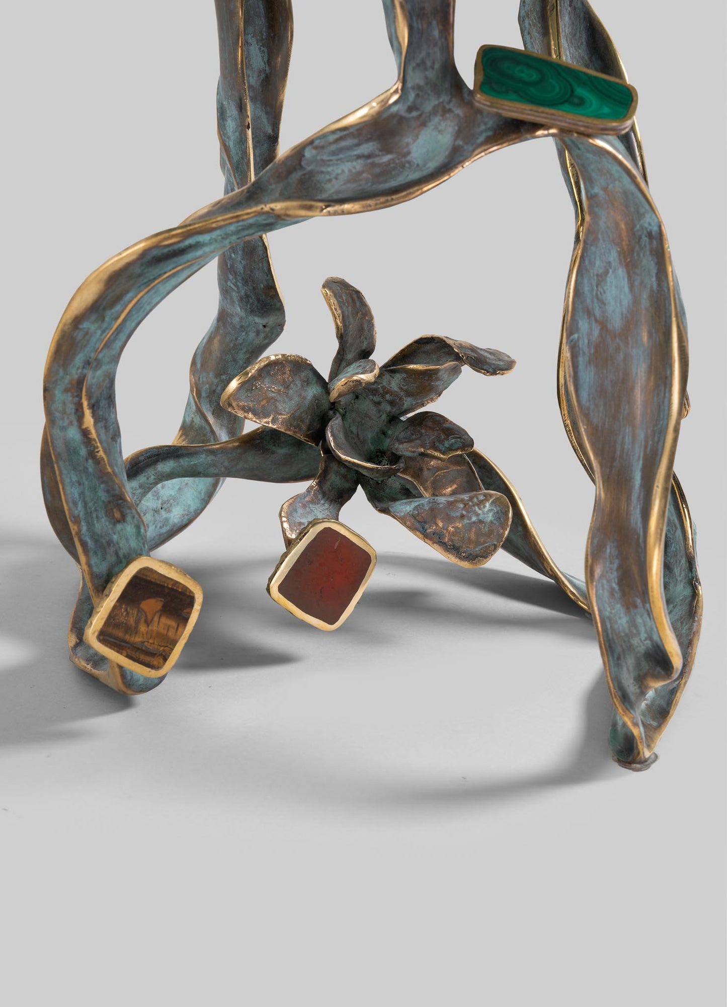 A sculptural decorative object made of metal with a distressed, aged finish and gold accents, featuring organic, flowing shapes and embedded colored stones or glass pieces in green, red, and amber tones.