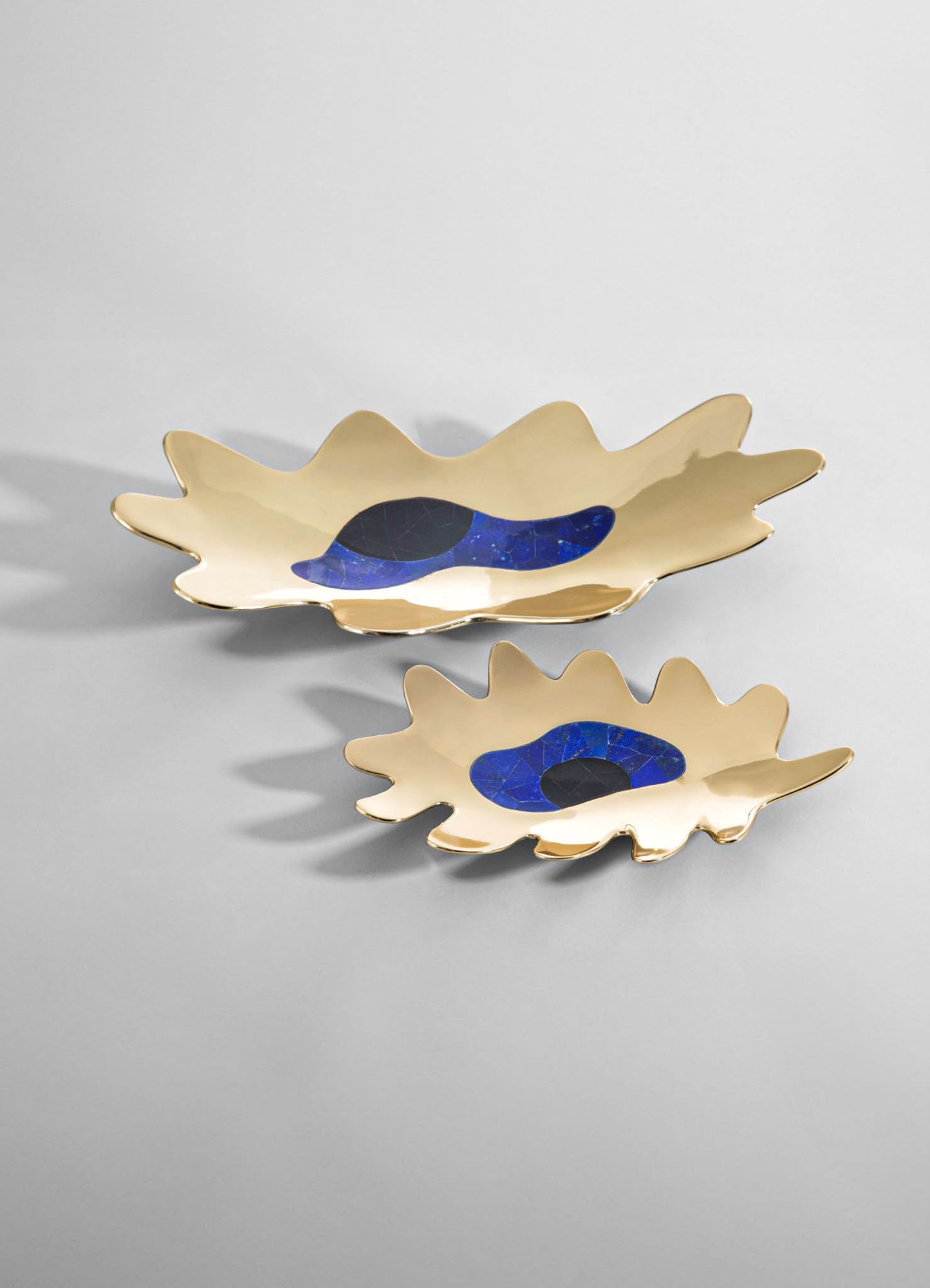 Ceramic or metal decorative trays with a scalloped edge design in a neutral beige tone, featuring abstract blue and black motifs in the center. The trays have a glossy finish and a modern, artisanal style.