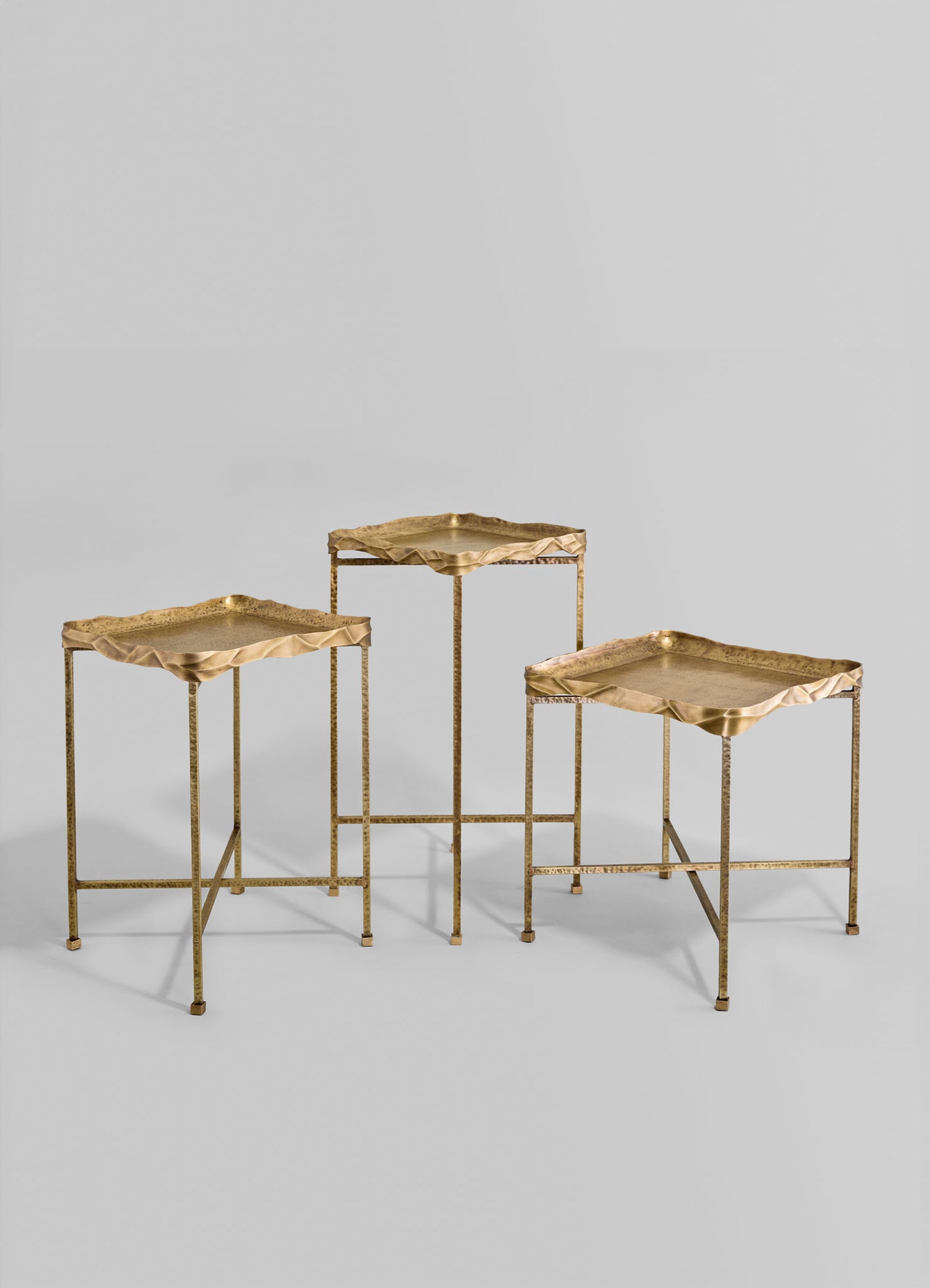 Three metal side tables with irregular, wavy-edged brass tops and slender, textured brass legs arranged against a plain background. The tables feature a handcrafted style with a warm, metallic gold finish.