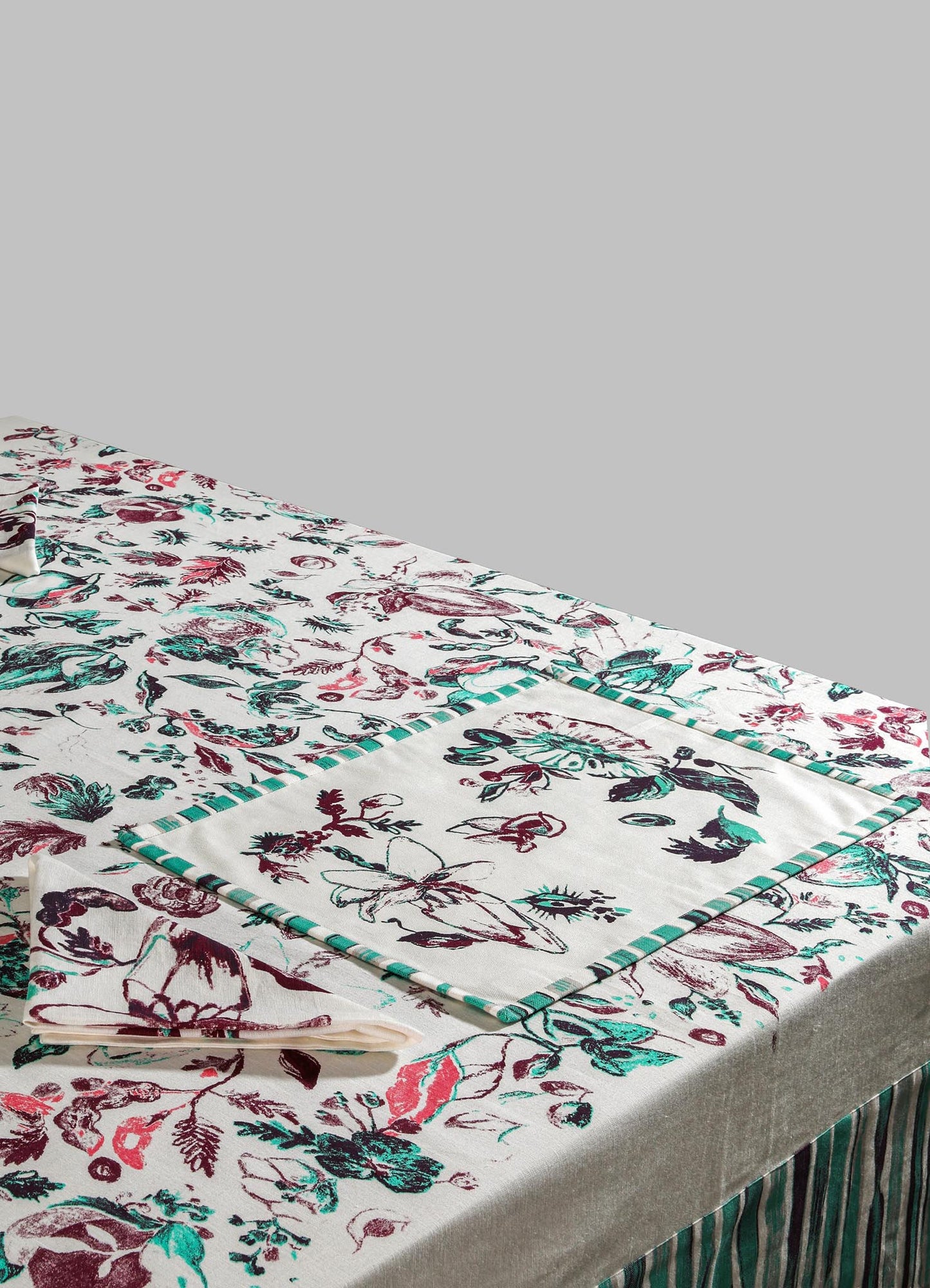 Cotton tablecloth with a botanical and floral print in shades of red, teal, and black, featuring a striped border detail. The fabric has a smooth, matte finish and a neutral beige base, creating a contemporary and elegant look.