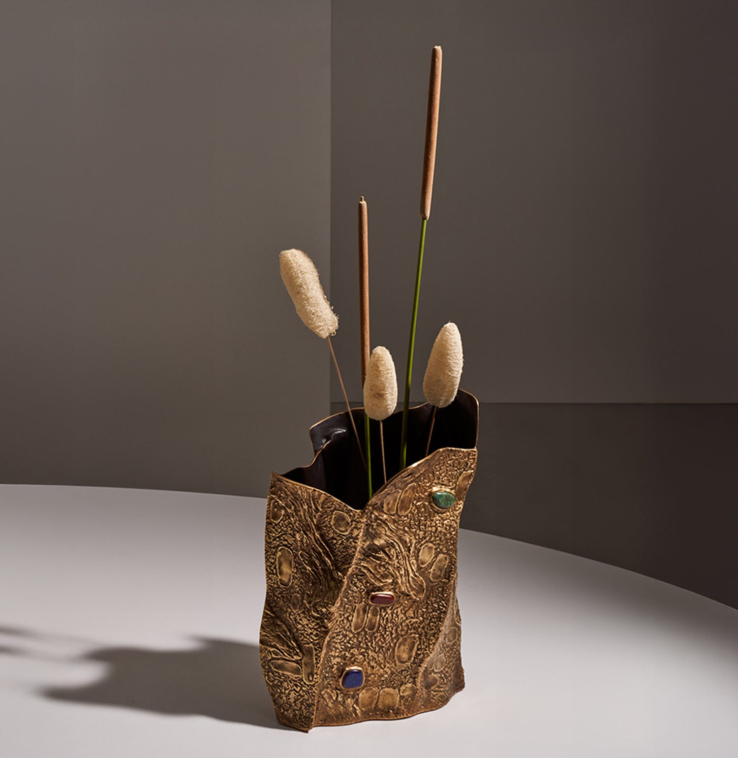 A decorative vase with a textured, metallic gold finish and embedded colorful stones, holding a arrangement of dried botanical elements, with a contemporary design style.