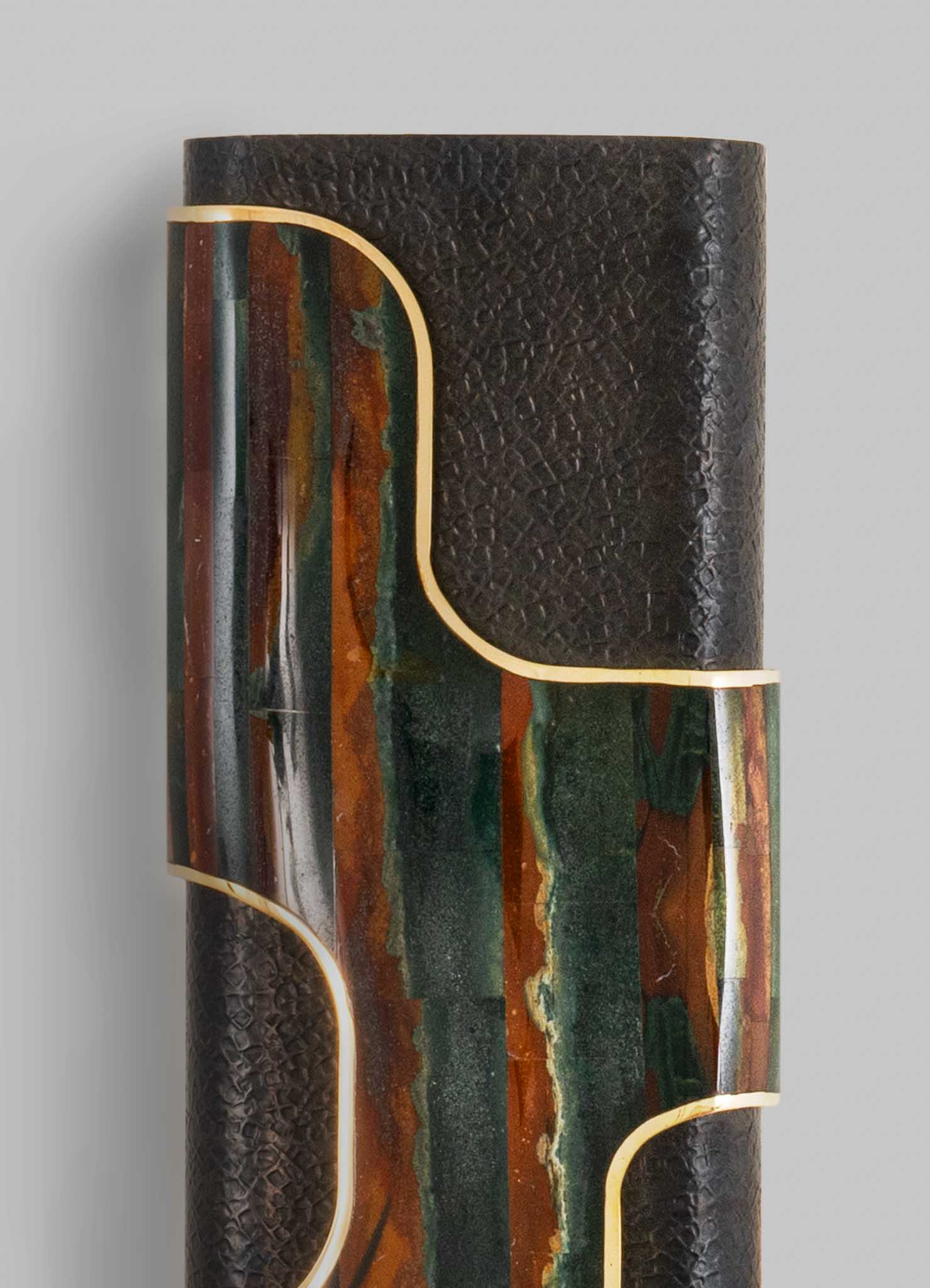 A tall cylindrical lamp with a textured black exterior and a multicolored, abstract-patterned inlay featuring earthy tones like green, orange, and brown, accented with gold trim.