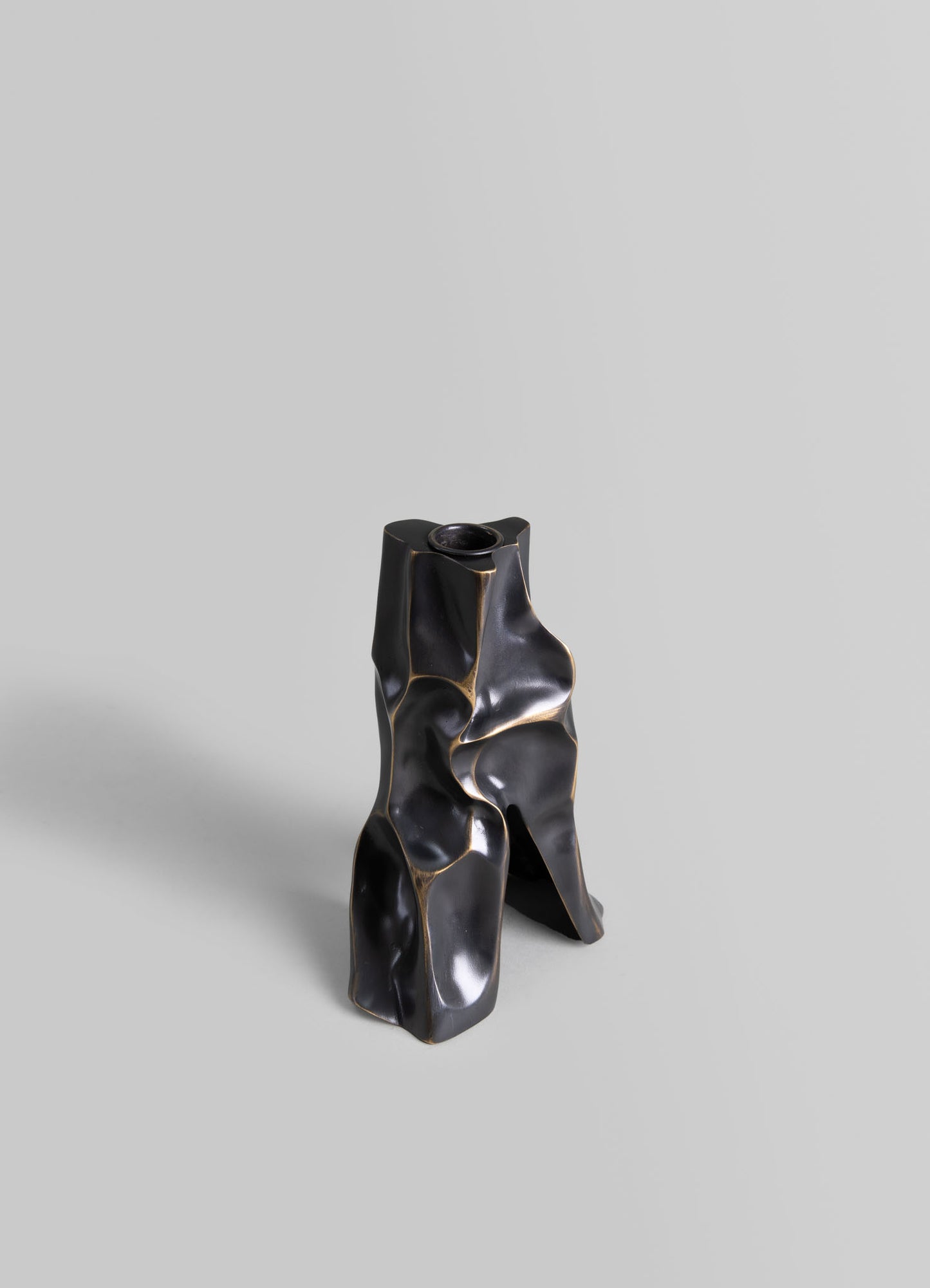 A sculptural vase made of dark, polished ceramic or metal with irregular, organic forms and flowing curves, set against a plain light gray background.