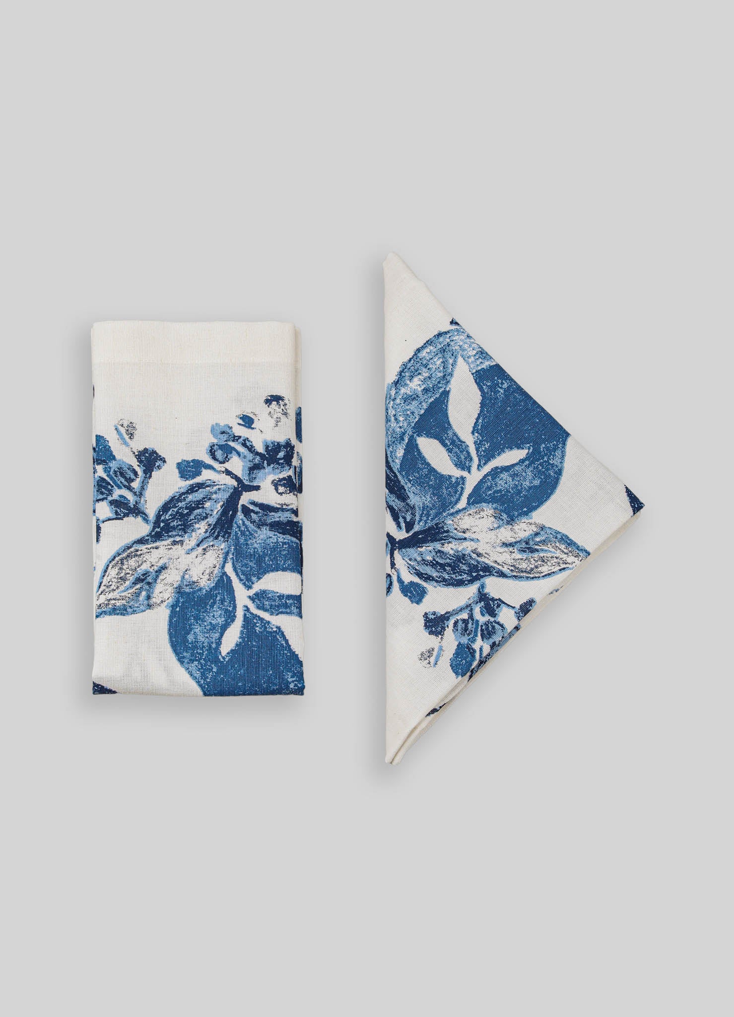Cotton napkins with a blue botanical print featuring leaves and flowers, one folded and the other partially unfolded. The fabric has a crisp, smooth texture with a contemporary design.