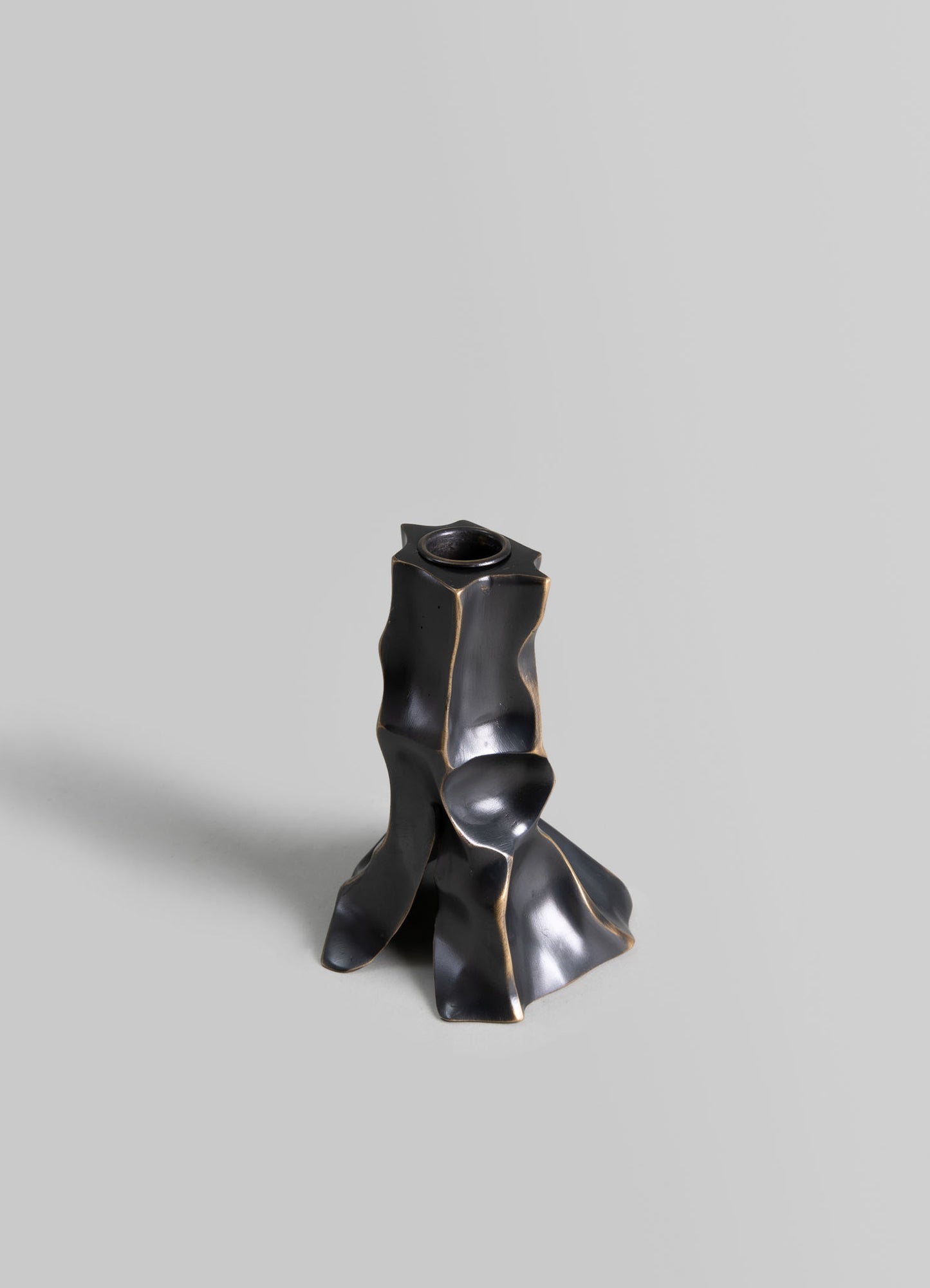 A sculptural vessel or vase in matte black material with irregular, organic contours and subtle metallic accents, displayed on a plain, light gray background.