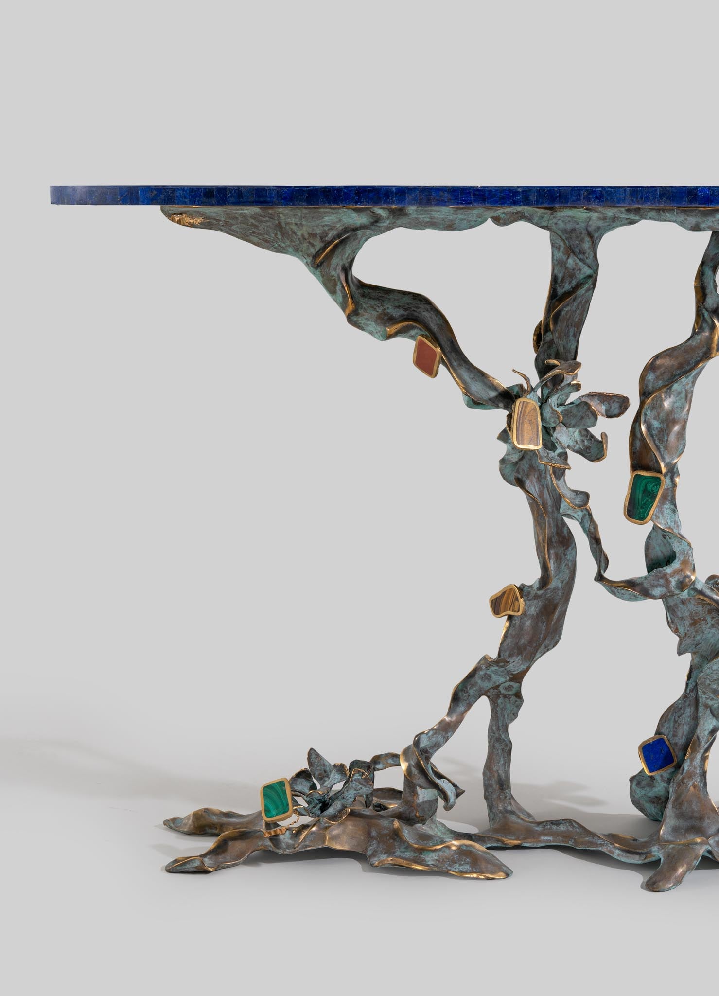 A metal table with an irregular organic shape, featuring a sculptural base resembling intertwined branches, finished in a dark patina with gold accents. The table top is a smooth, dark blue material that contrasts with the textured base.