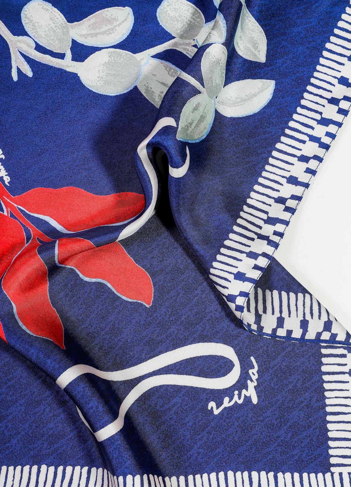 Printed silk scarf with a floral and geometric motif in shades of red, white, and blue, featuring a border with stripes and checkered patterns. The fabric has a smooth, glossy finish.