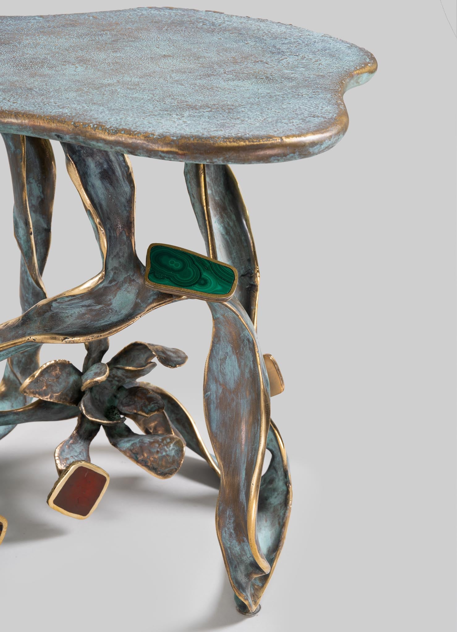 A decorative table with a textured, irregularly shaped top in a muted, metallic finish and sculptural metal legs resembling plant stems with leaf motifs, accented by gold trim. It features green and reddish stones set into the base, contributing to an artistic, handcrafted style.