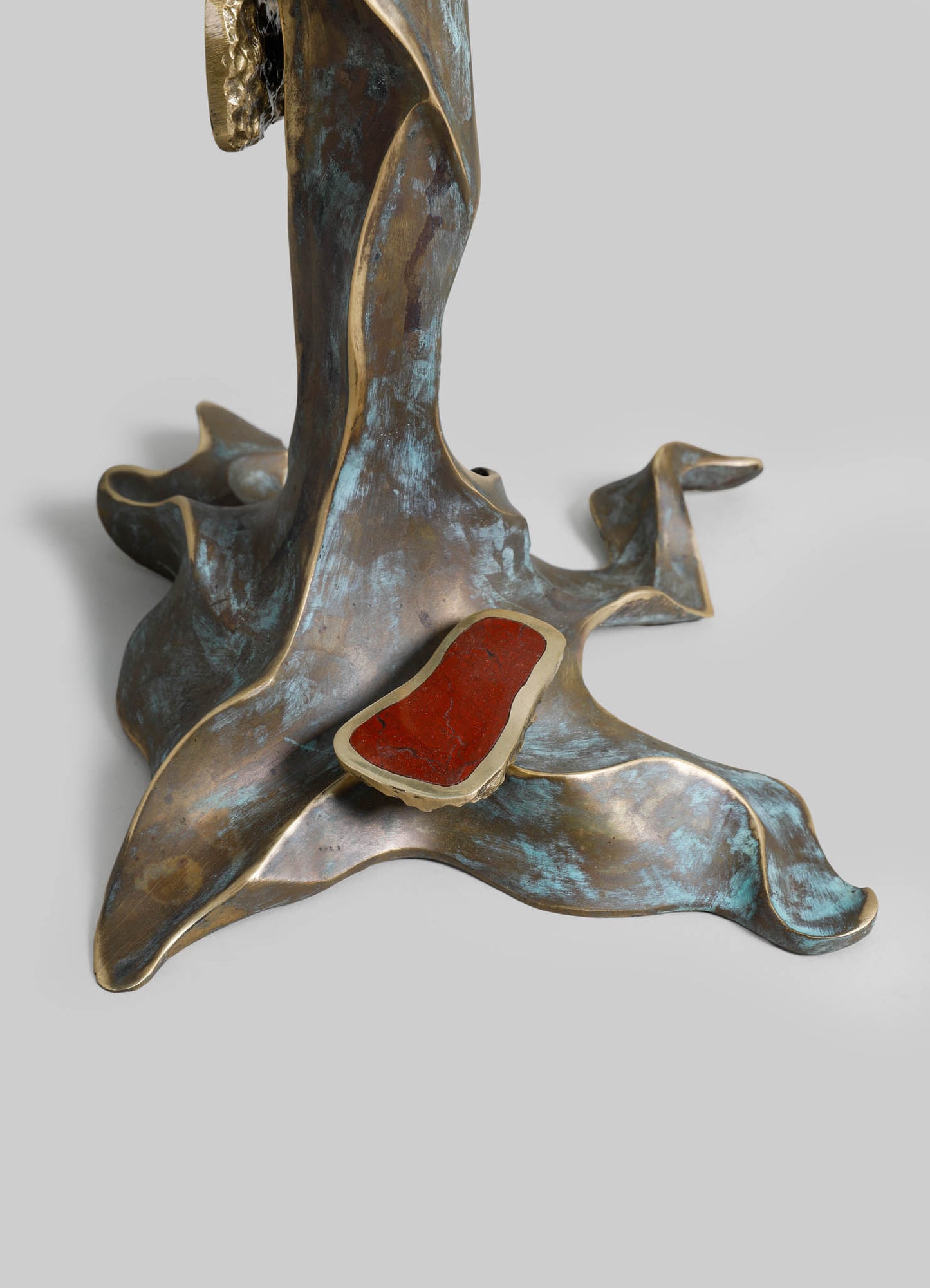 A sculptural base with an organic, flowing form made of metal with a distressed, patinated finish in shades of blue and bronze, featuring a red stone accent on its surface.