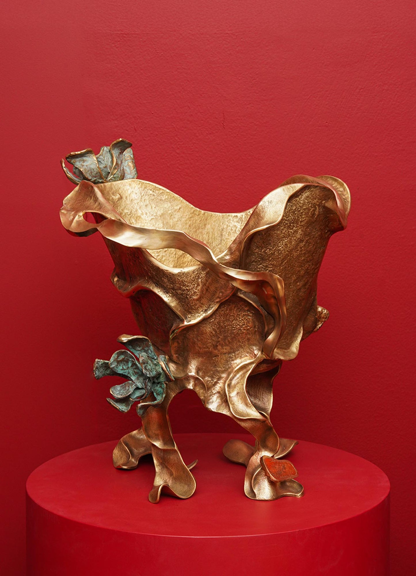 A sculptural decorative bowl made of metal with an organic, flowing form and a mix of bronze and patinated blue-green finishes, displayed on a red round pedestal against a red wall.
