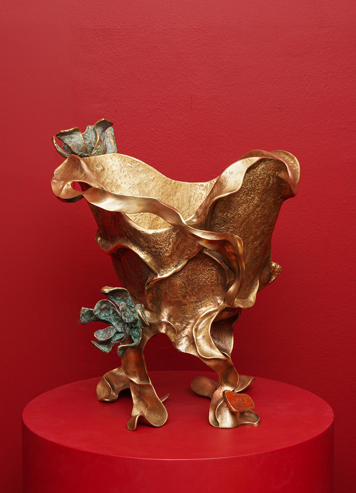 A sculptural decorative bowl made of metal with an organic, flowing form and a mix of bronze and patinated blue-green finishes, displayed on a red round pedestal against a red wall.