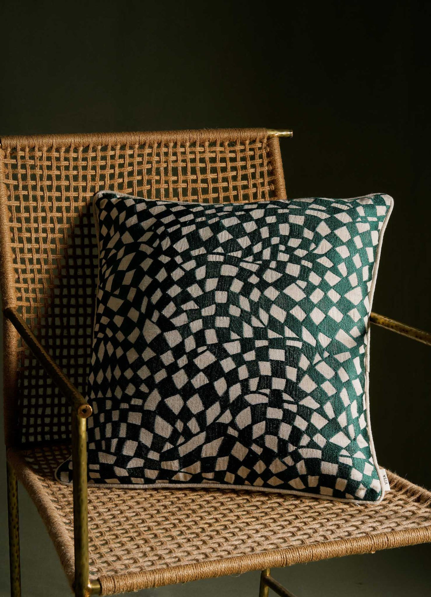 A decorative cushion with a black and white geometric pattern, placed on a woven rattan chair with brass armrests, against a dark background. The cushion appears to be made of fabric with a contemporary design motif.
