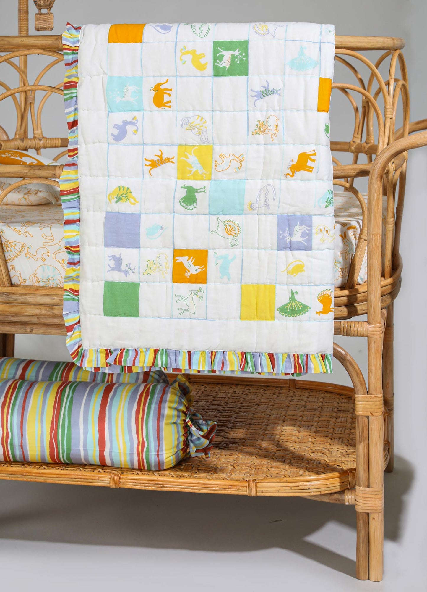 Colorful patchwork quilt with animal embroidery featuring dinosaurs in bright yellow, green, blue, and orange hues, bordered with striped multicolor trim. The quilt is draped over a rattan baby crib alongside a matching striped bolster pillow, creating a playful and casual nursery decor.