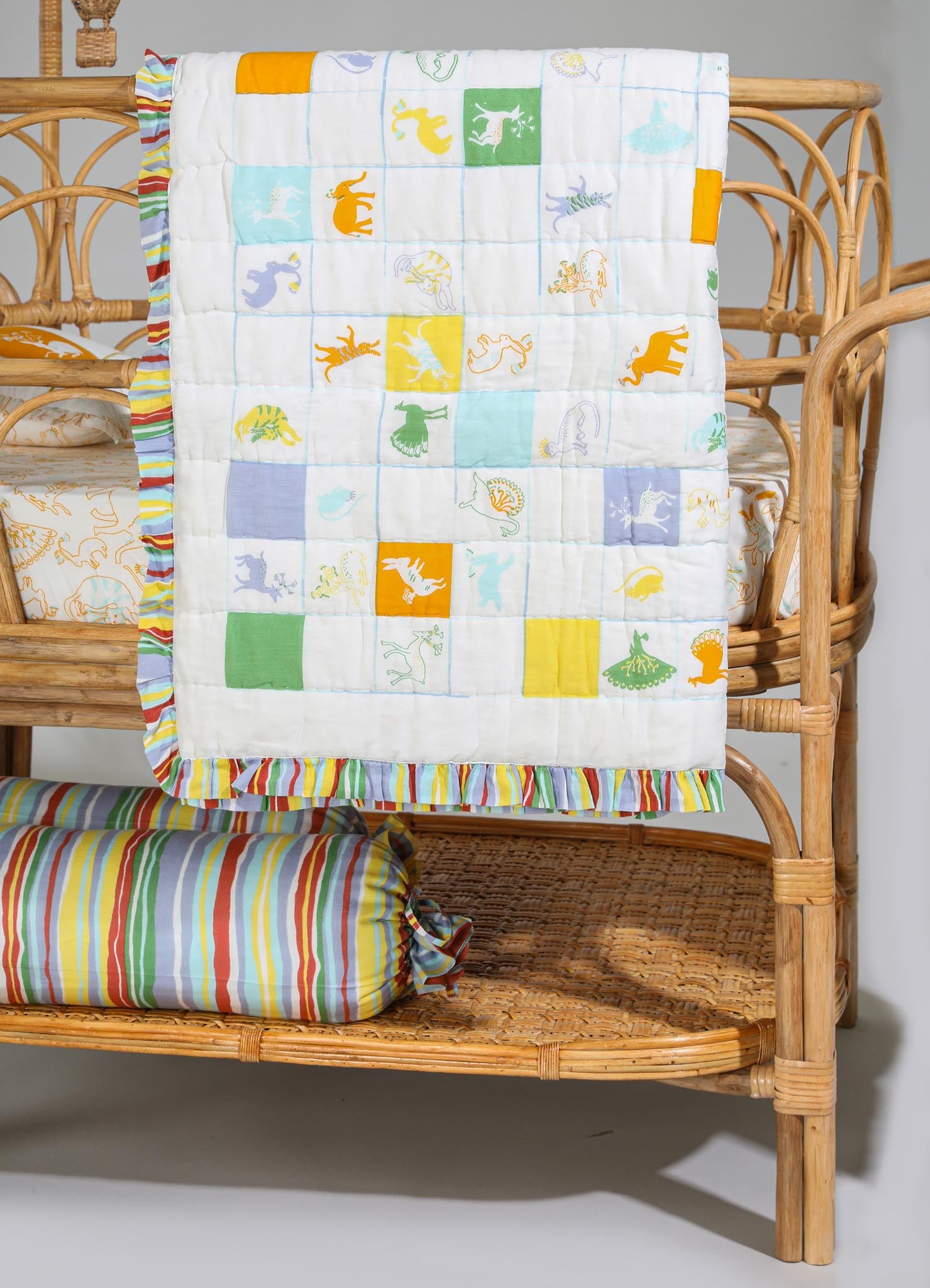 Colorful patchwork quilt with animal embroidery featuring dinosaurs in bright yellow, green, blue, and orange hues, bordered with striped multicolor trim. The quilt is draped over a rattan baby crib alongside a matching striped bolster pillow, creating a playful and casual nursery decor.