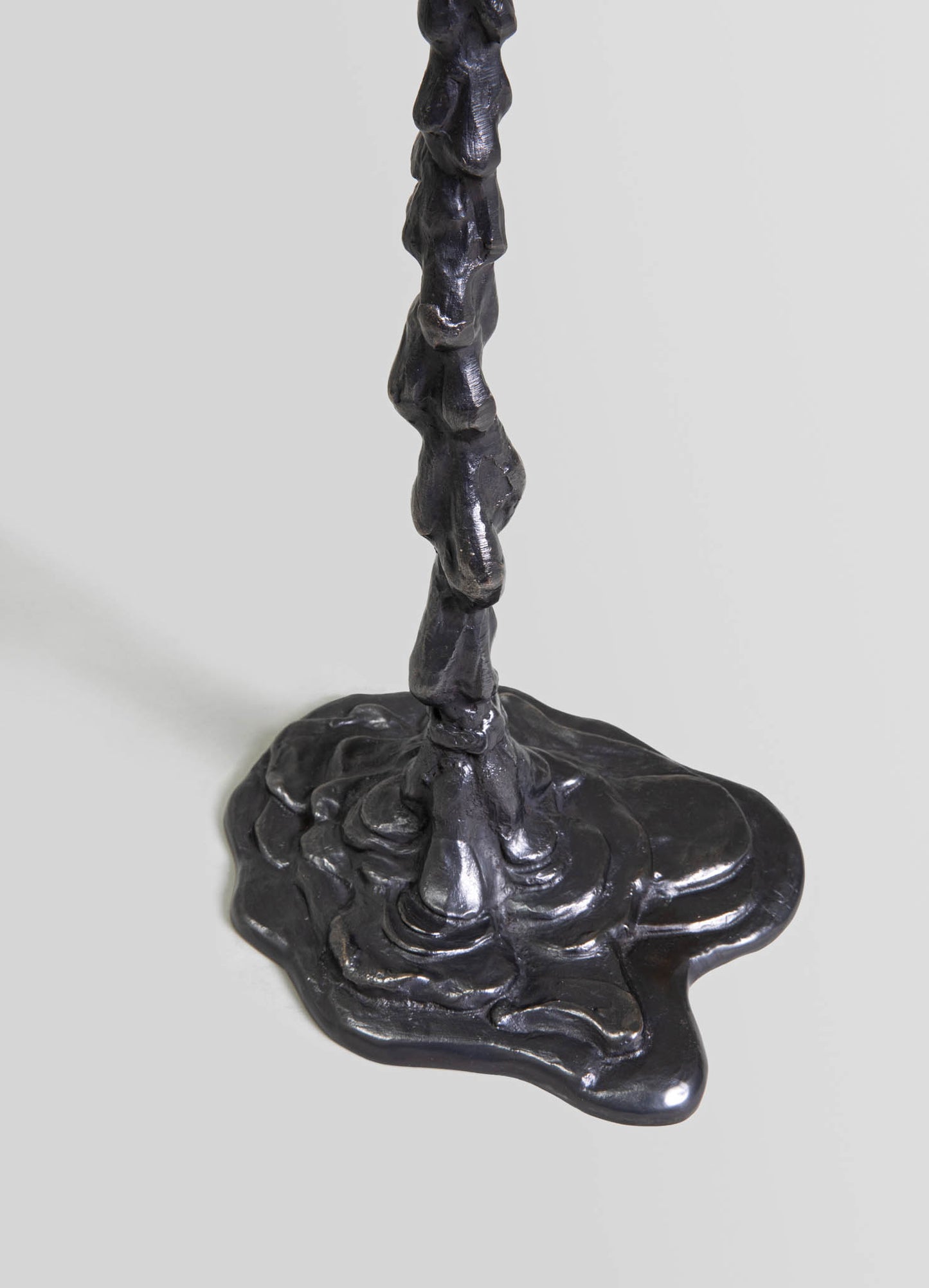 A black metal candlestick holder with a textured, organic design featuring a tall, irregularly shaped stem and a wide, uneven base. The finish is matte with a slightly reflective surface.