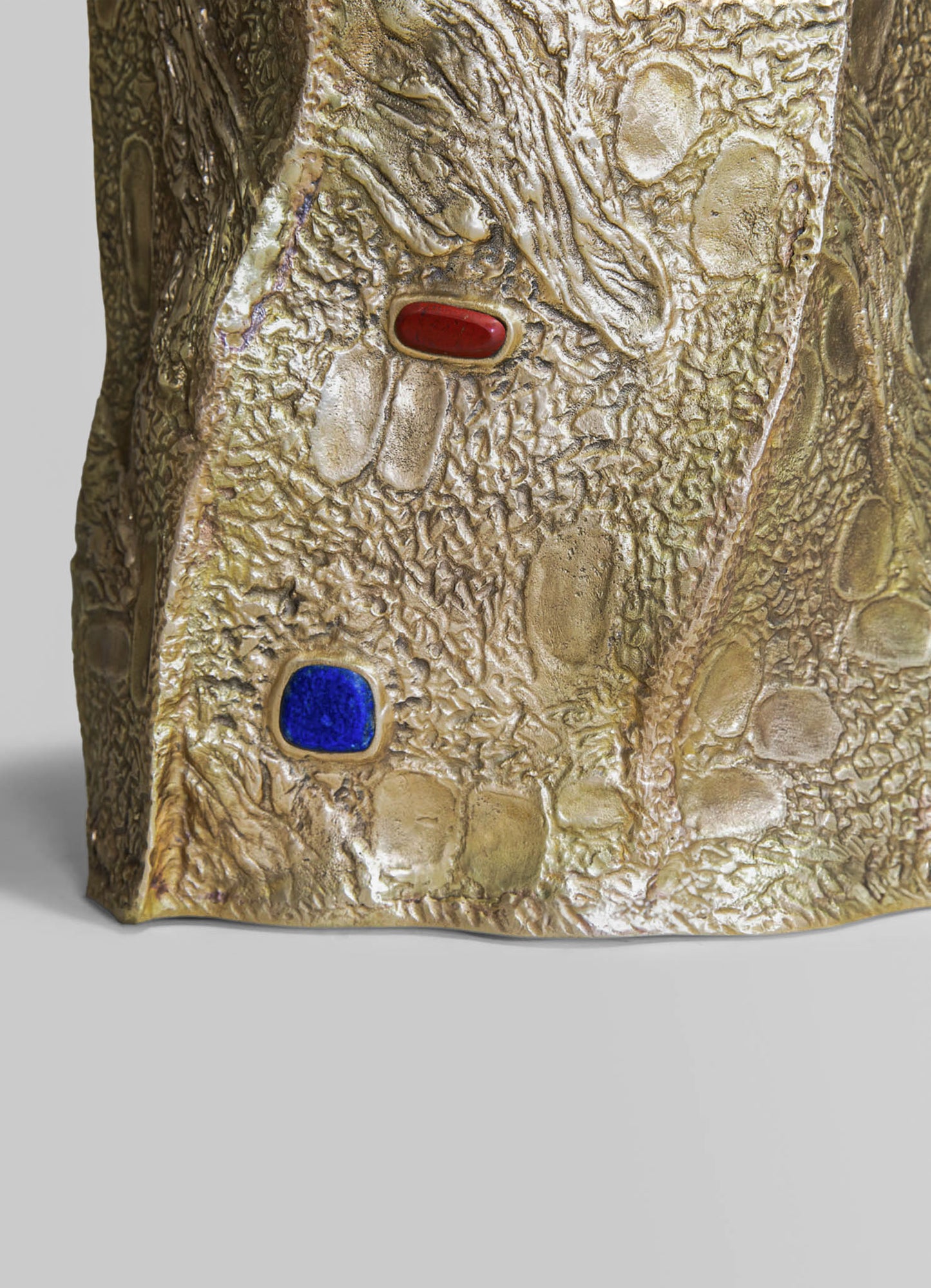 Close-up of a textured sculpture or decorative object with a rough, organic surface in beige and gold tones, featuring embedded geometric shapes in red and blue. The piece has an artisanal, handcrafted style with a focus on natural textures and bold, colorful accents.