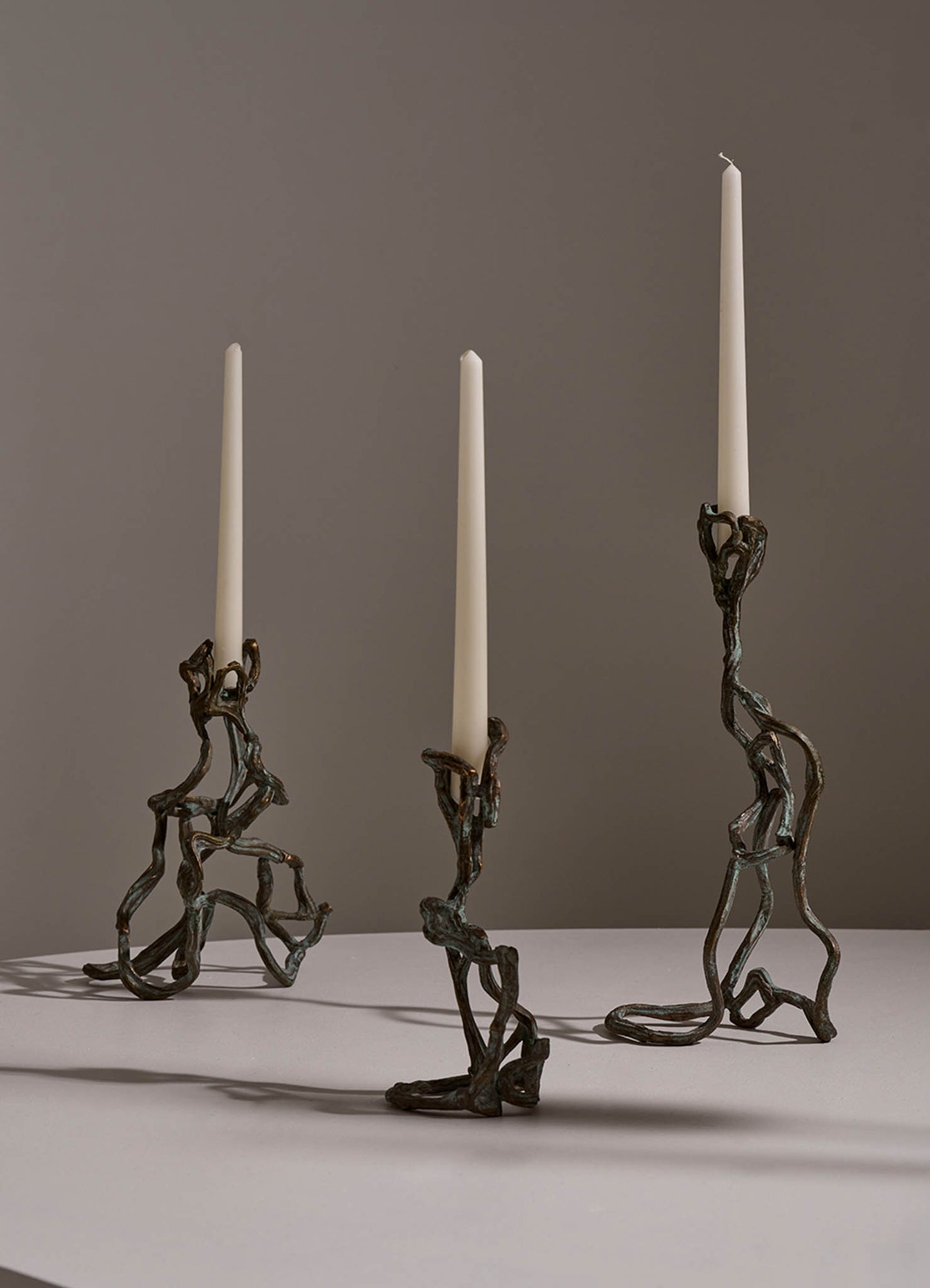 Three candlesticks with irregular, twisted bronze metal bases and tall white taper candles, set against a plain background.
