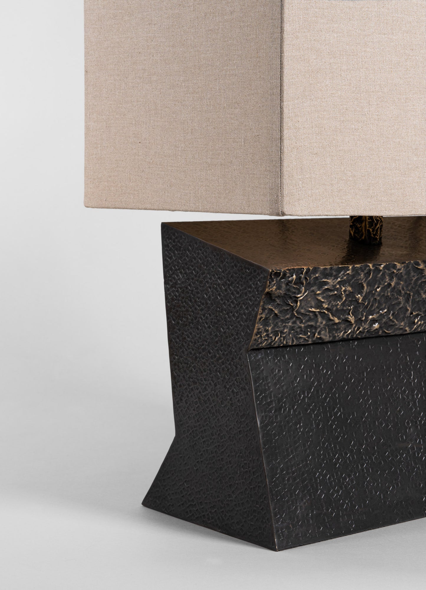 A close-up of a table lamp featuring a beige fabric shade with a smooth texture and a black base with a textured, geometric pattern resembling carved or embossed metal or leather. The base has an angular, modern design with a matte finish, creating a contrast with the soft, natural tone of the lampshade.