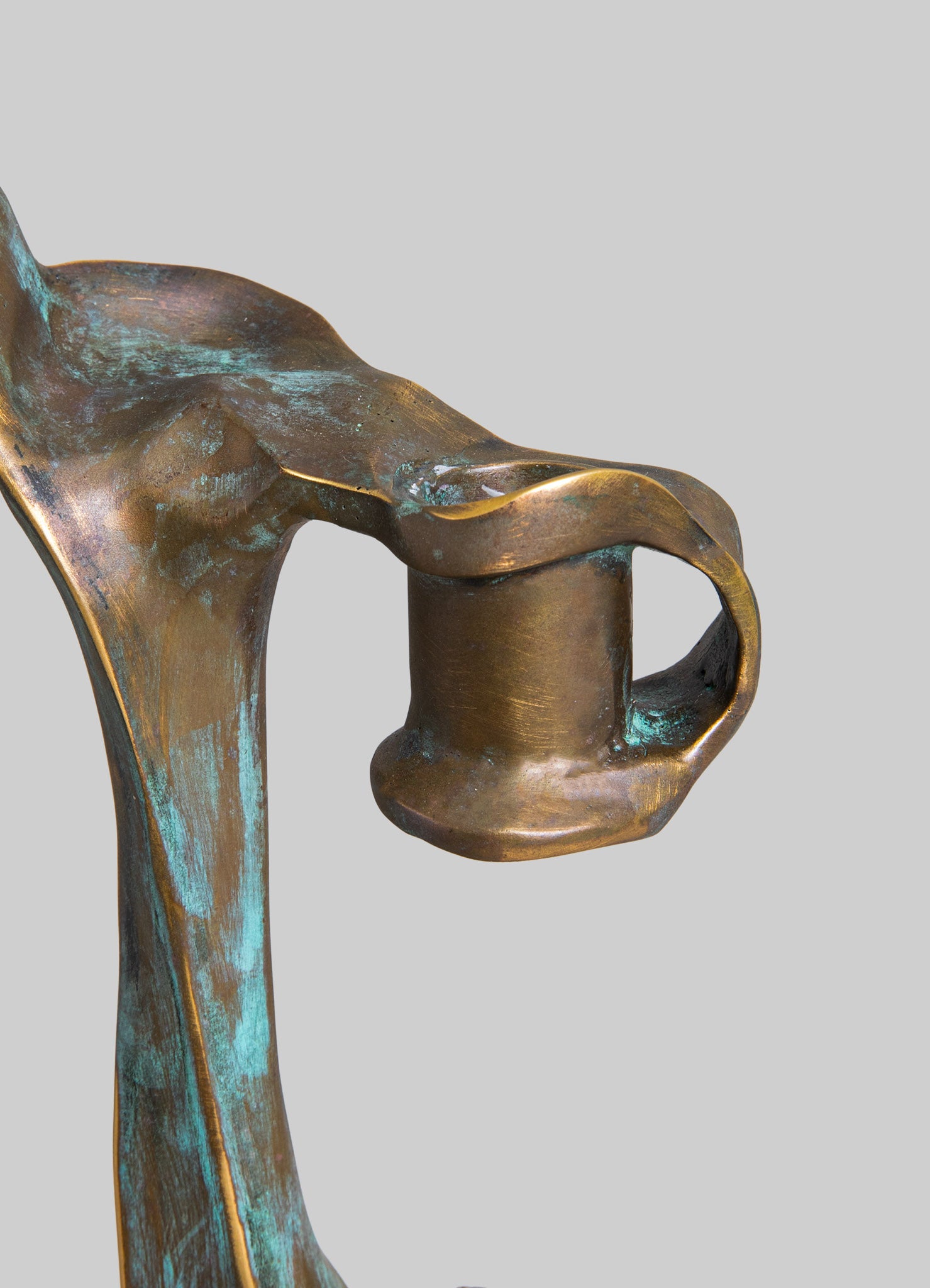 Close-up of a decorative metal sculpture with a weathered bronze finish and patina effects, featuring smooth, flowing contours and organic forms with hints of turquoise and green hues. The sculpture exhibits handcrafted artistry and an abstract, contemporary style.