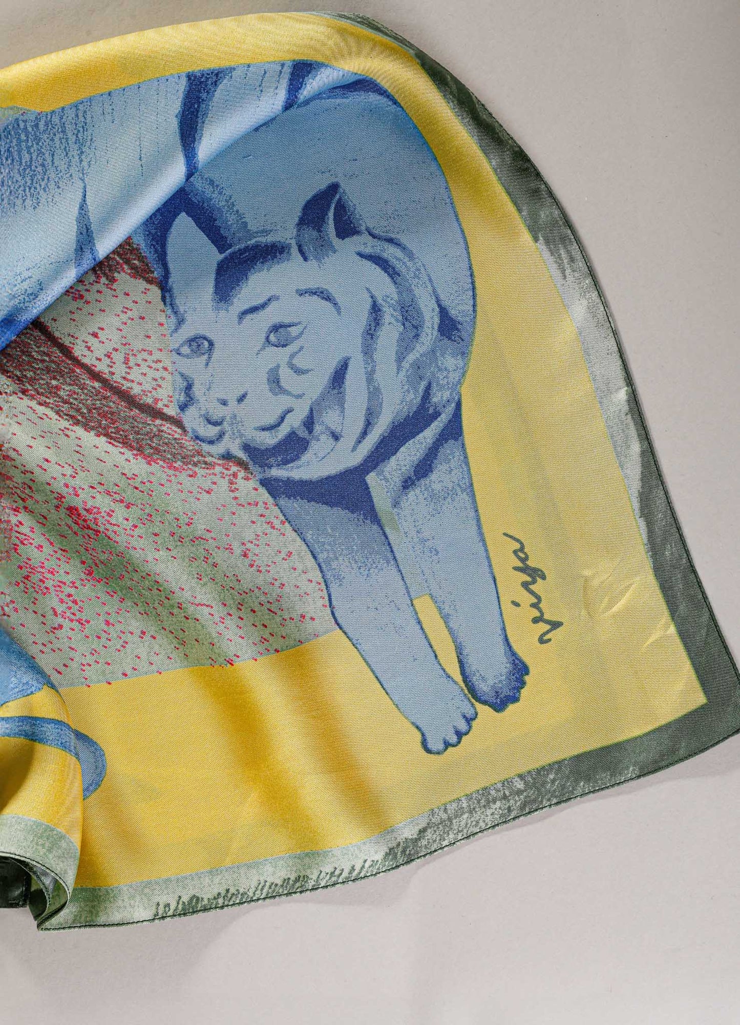 Silk scarf featuring a colorful portrait of a lion with a mane, in shades of blue, yellow, and red, with signature detail in black. The fabric has a smooth, glossy finish with a vibrant, artistic design.