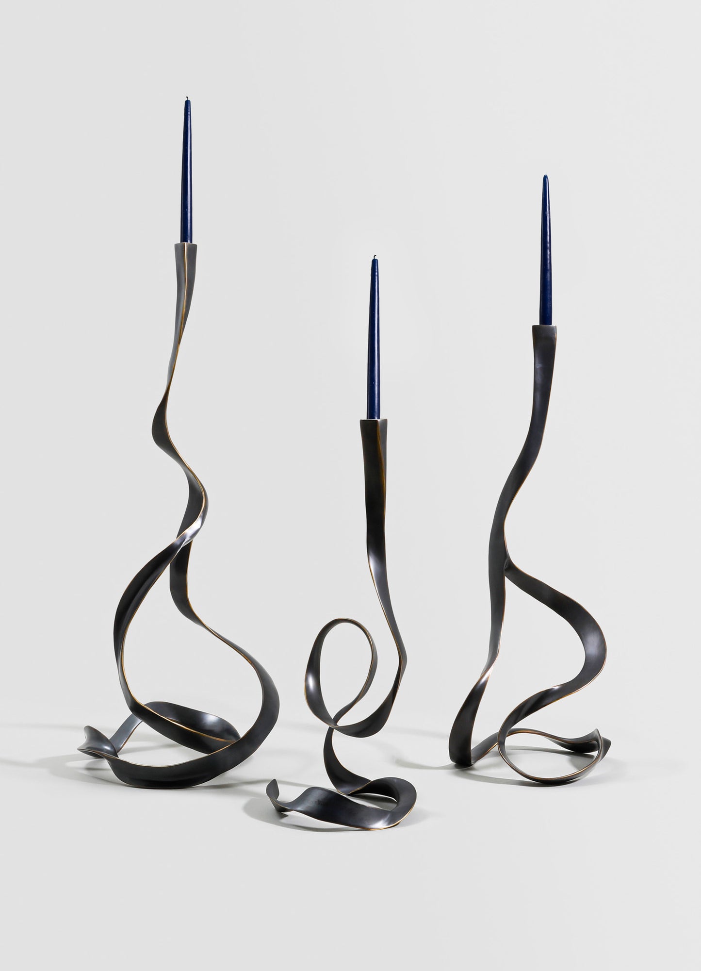 Three sculptural candle holders with twisting metal arms in matte black finish, each holding a slim, tapered blue candle against a plain background.