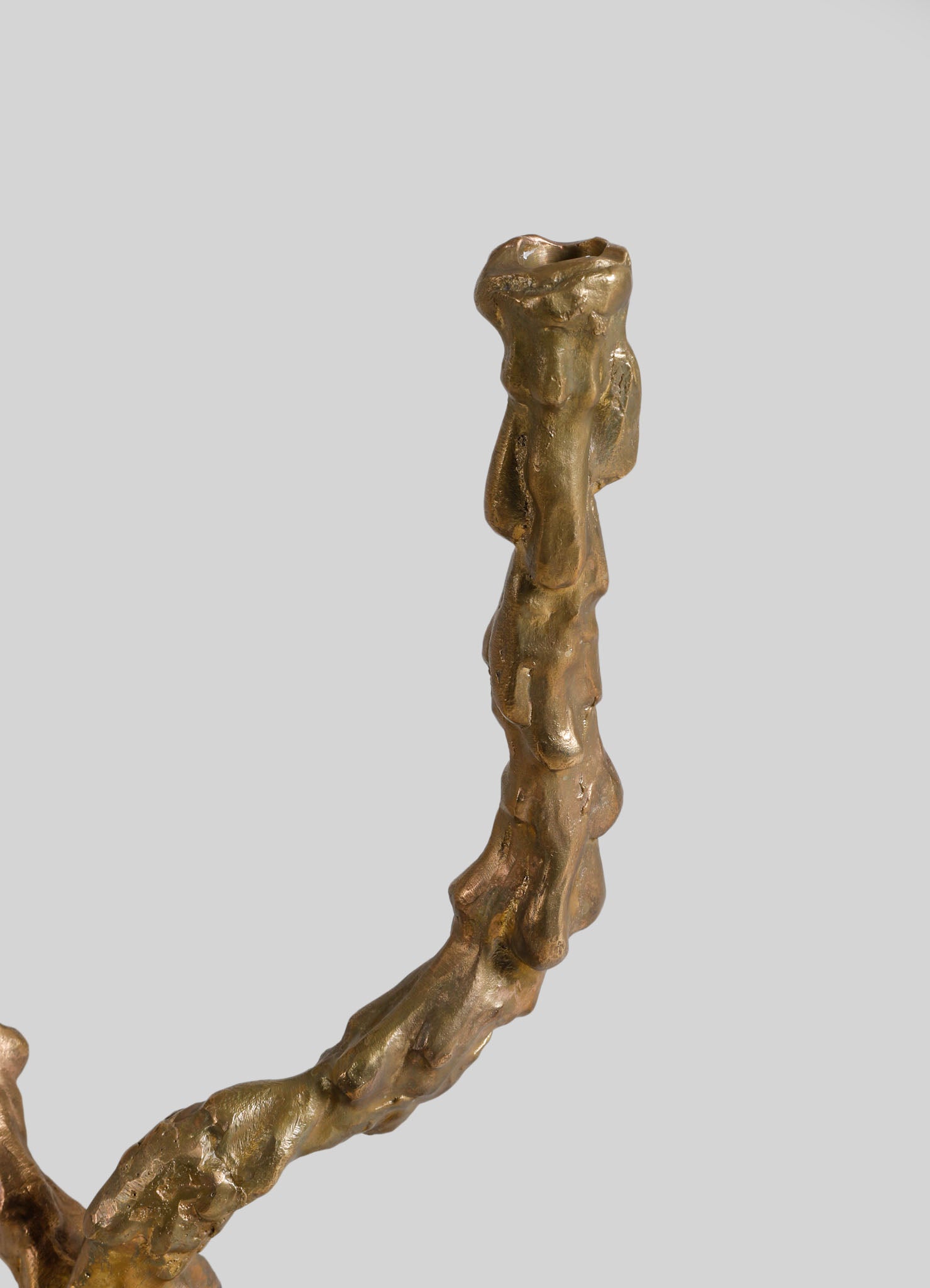A close-up of a sculptural candlestick holder with an organic, irregular shape, made of metallic material with a gold-tone finish. The design features a textured surface with flowing, natural contours, set against a plain gray background.