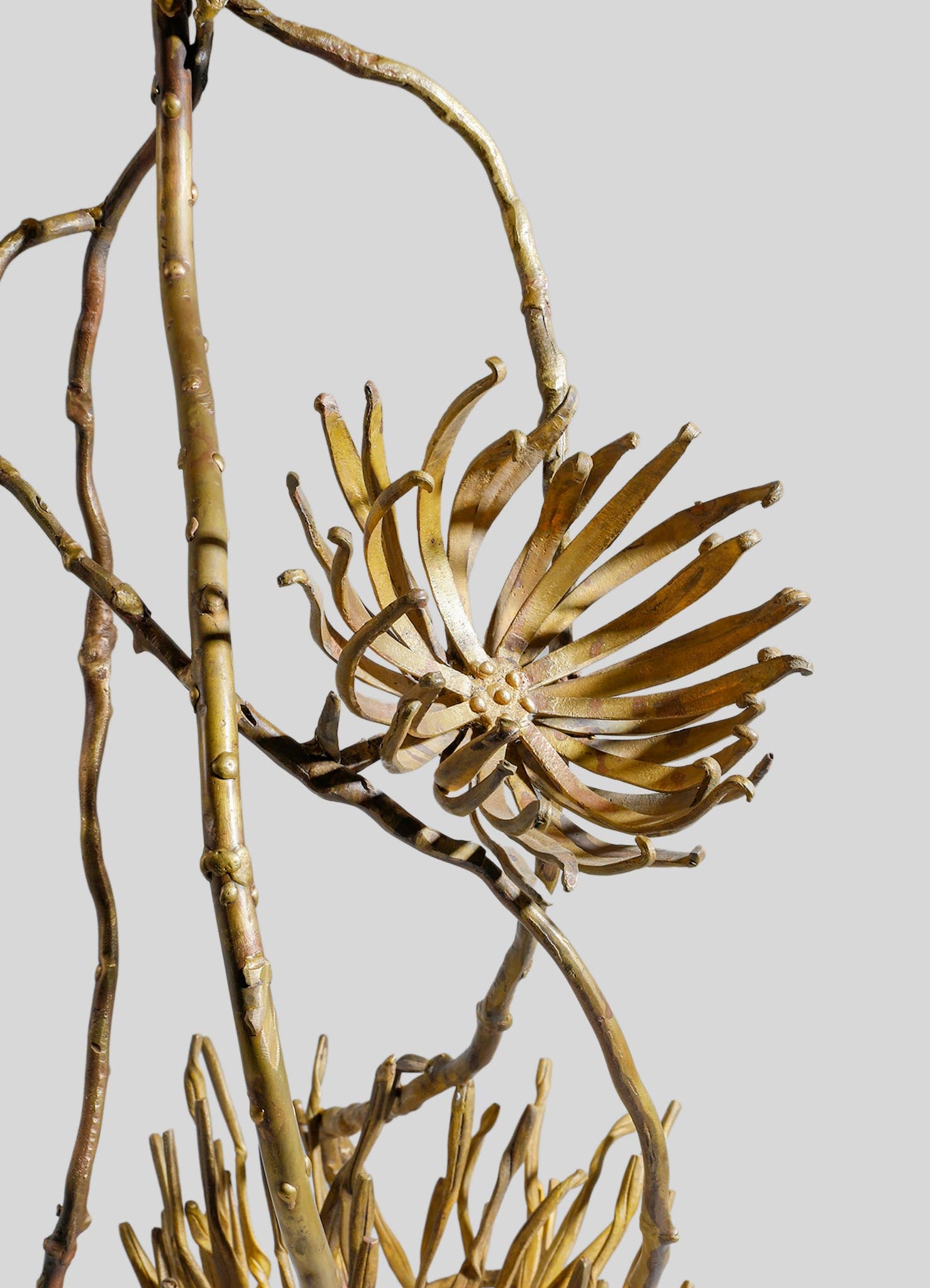 Golden metal sculpture resembling a floral or botanical motif with elongated, curved petals or leaves and a textured stem. The design has an elegant, handcrafted style with intricate detailing and a metallic finish.