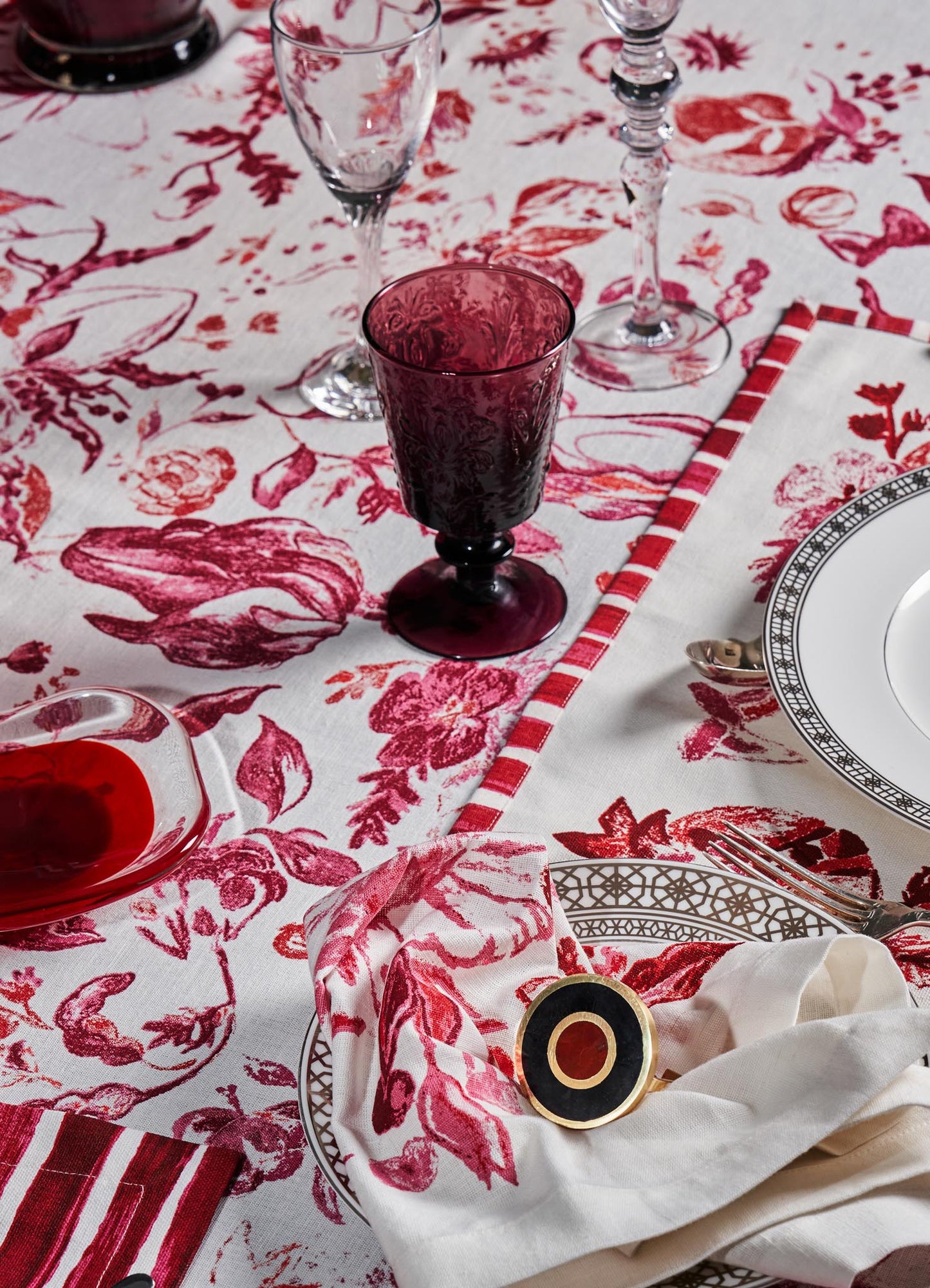 A table setting with fine glassware and dishware, including a clear glass wine glass, an ornate white plate with black geometric border, and a deep red goblet with floral pattern. The tablecloth features a white background with large red floral and botanical motifs, complemented by a matching cloth napkin with red and white stripes.