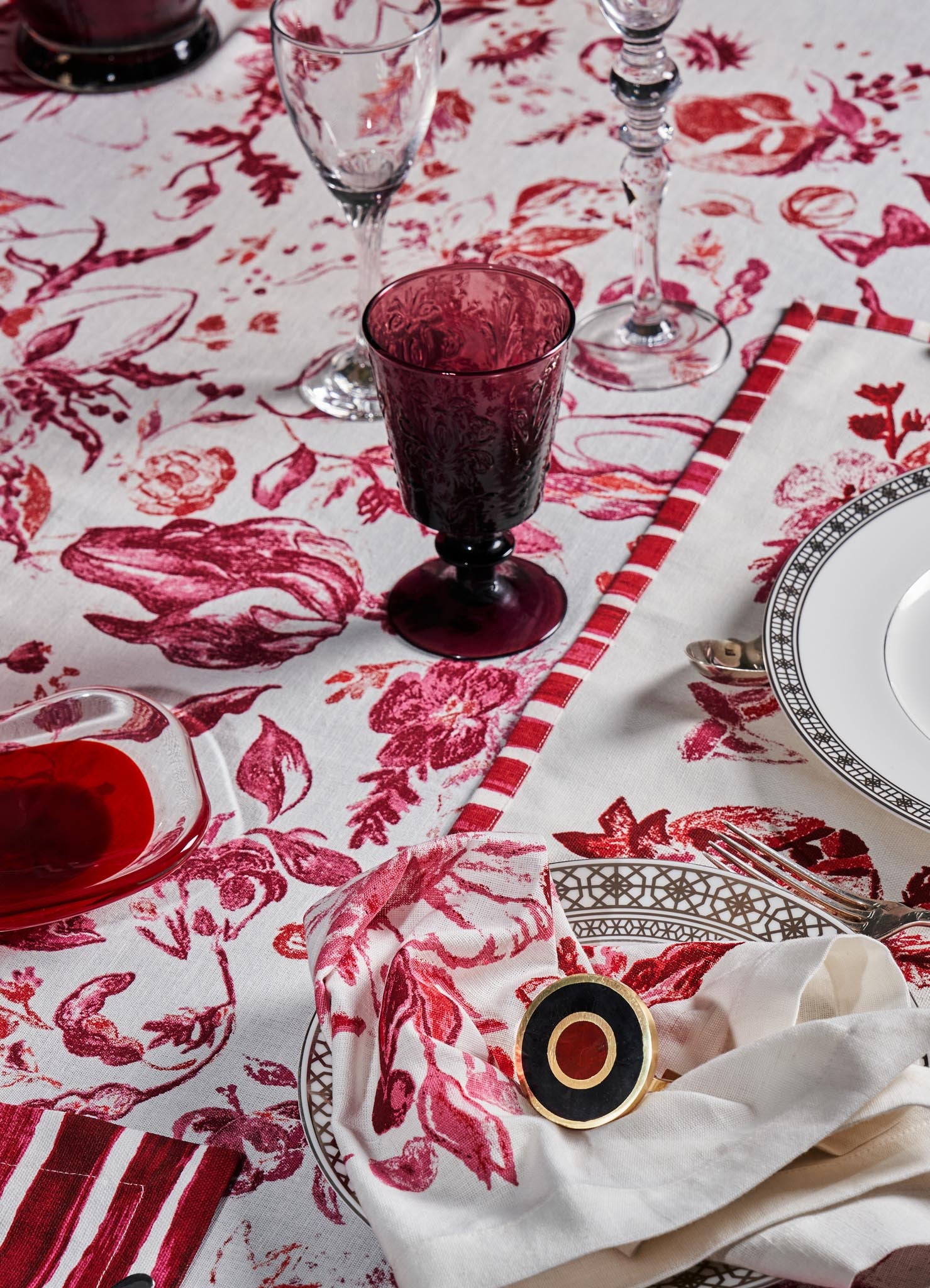 A table setting with fine glassware and dishware, including a clear glass wine glass, an ornate white plate with black geometric border, and a deep red goblet with floral pattern. The tablecloth features a white background with large red floral and botanical motifs, complemented by a matching cloth napkin with red and white stripes.