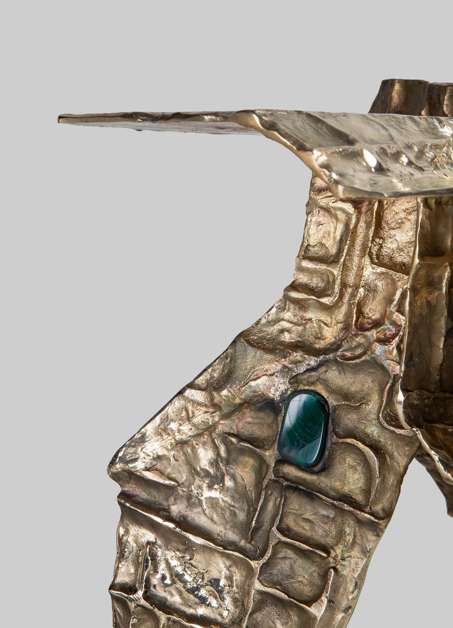 A metallic sculpture with a textured, rough finish in a gold-tone, featuring an angular, abstract form. The sculpture has an irregular surface with a polished, shiny appearance and a prominent blue-green stone embedded within the design.
