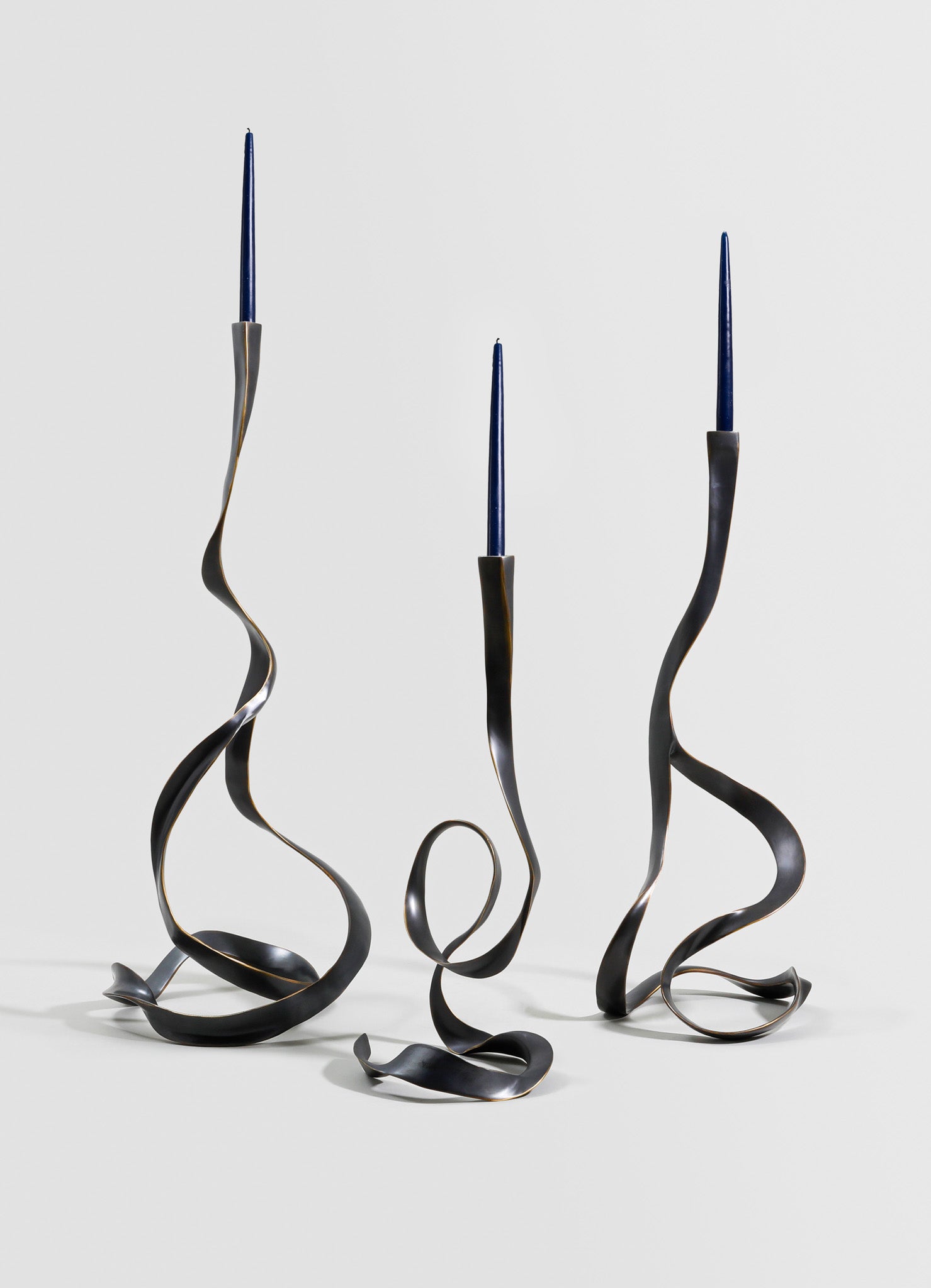 Three metal candlesticks with twisted, sculptural design and dark matte finish, each holding a tall, slender blue candle.