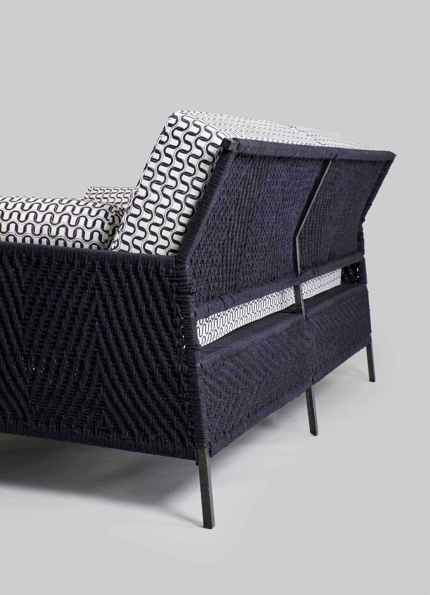 A modern armchair with a woven black rattan frame, featuring a patterned fabric cushion with black and white geometric designs and slim black metal legs.