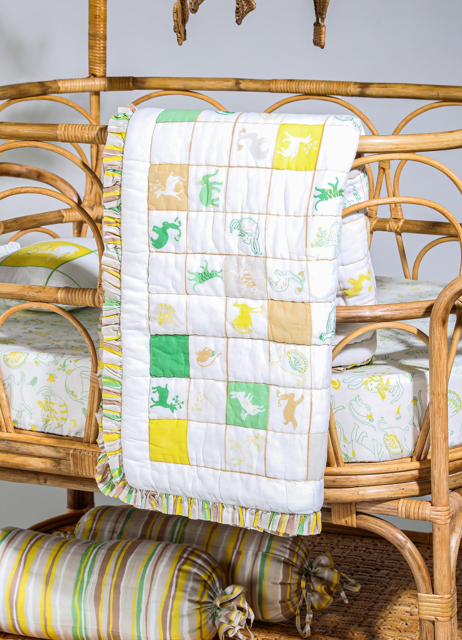 A quilted blanket featuring a playful animal and dinosaur print in yellow, green, and beige tones, with a ruffled trim along the edges. It drapes over a rattan crib, which has an organic, rustic style, complemented by matching bedding with similar printed motifs. A striped bolster pillow with yellow, white, and beige colors is also visible at the base of the crib.